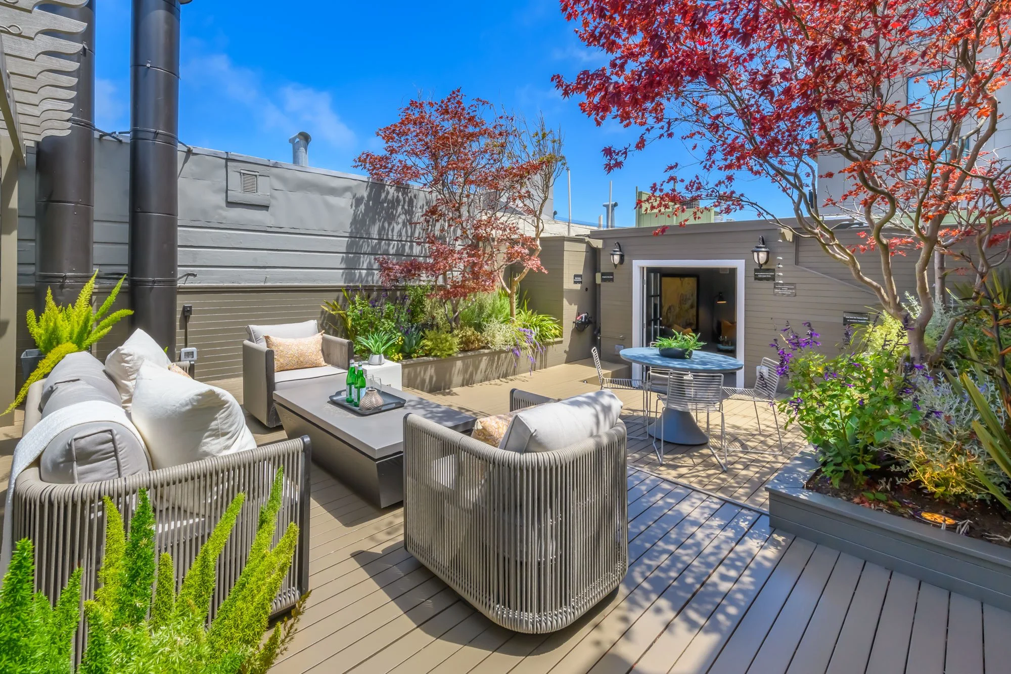 Outdoor patio area with beige and gray furniture, surrounded by potted plants and trees with red leaves, under a bright blue sky with some clouds.