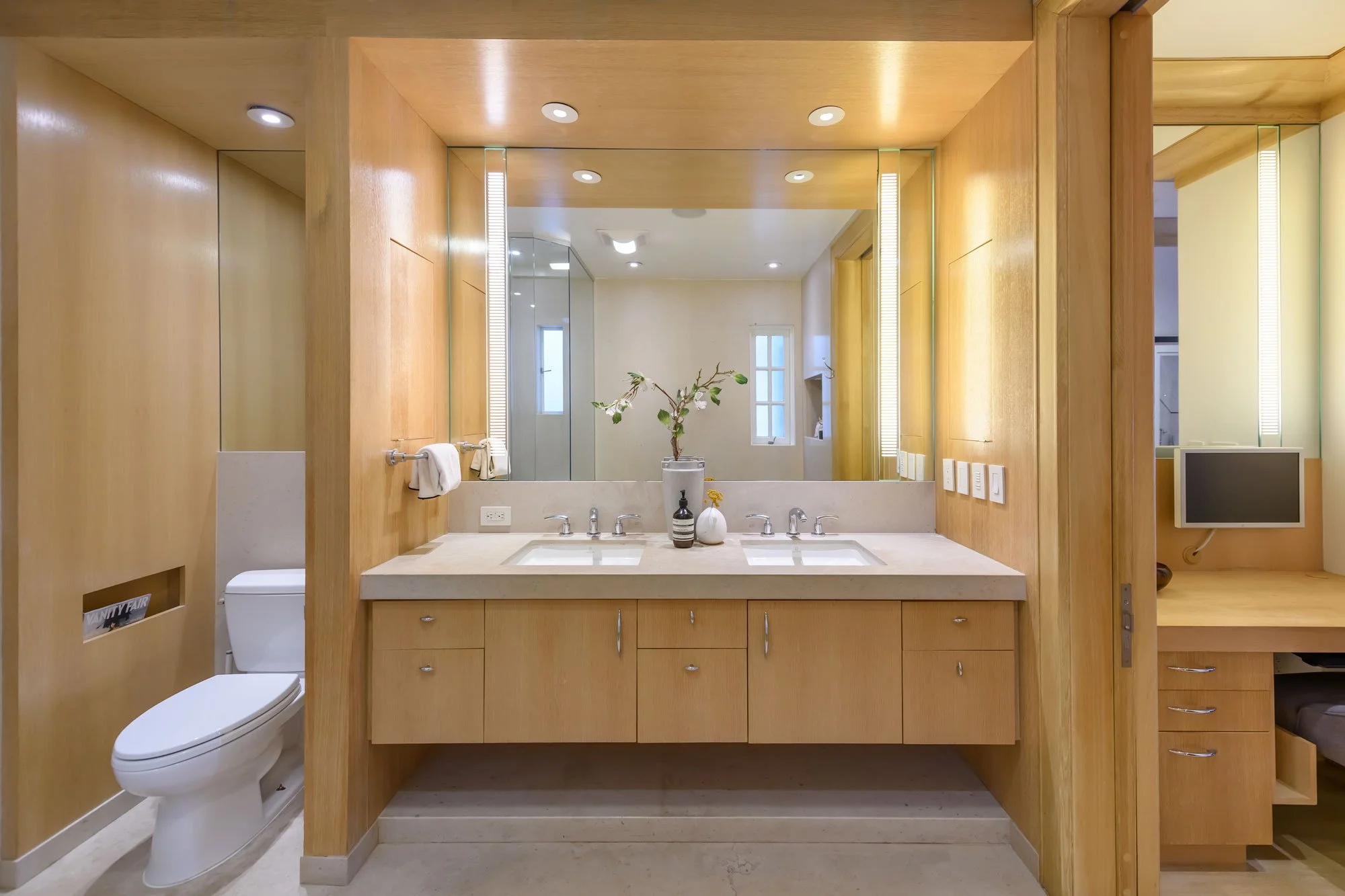 Modern bathroom with wooden accents, double sink vanity, large mirror, small window, decorative vases, and toilet visible to the left.