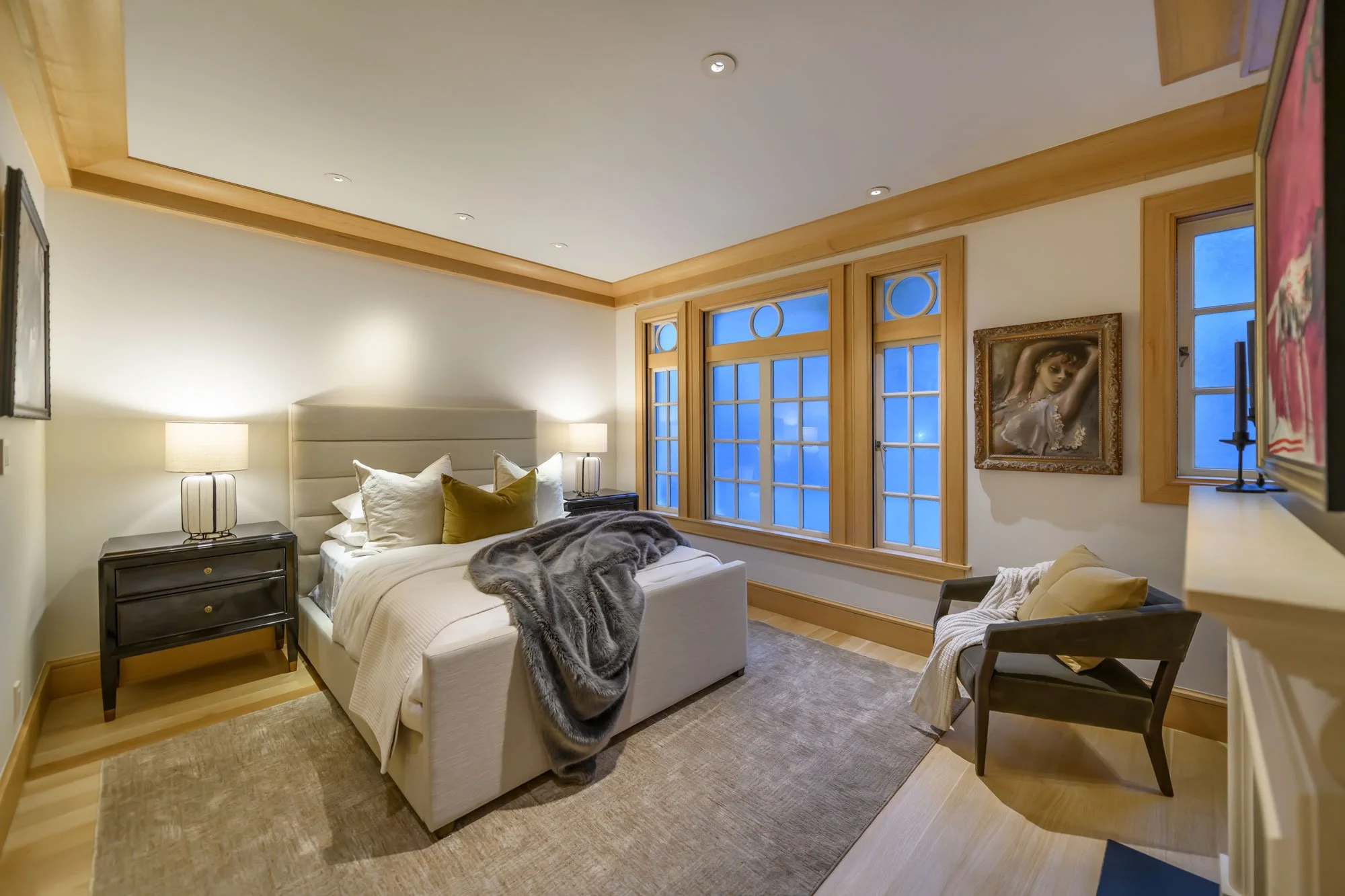 A cozy bedroom with a white bed, beige pillows, a gray blanket, and two black nightstands with lamps. Large windows with wooden frames and blue sky visible, framed artwork on the wall, light-colored wood flooring, and warm lighting.