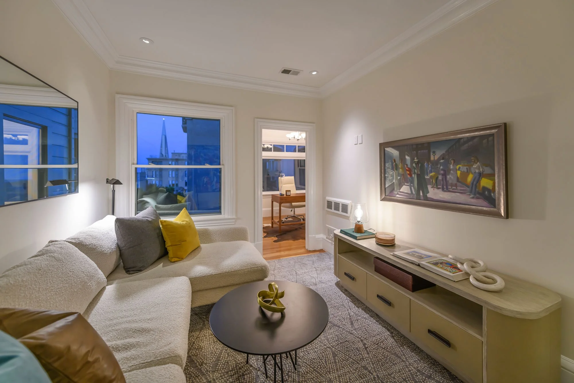 Living room with beige sofa, yellow and gray pillows, black round coffee table, white entertainment console, framed artwork on wall, two windows showing cityscape, and partial view into a home office.
