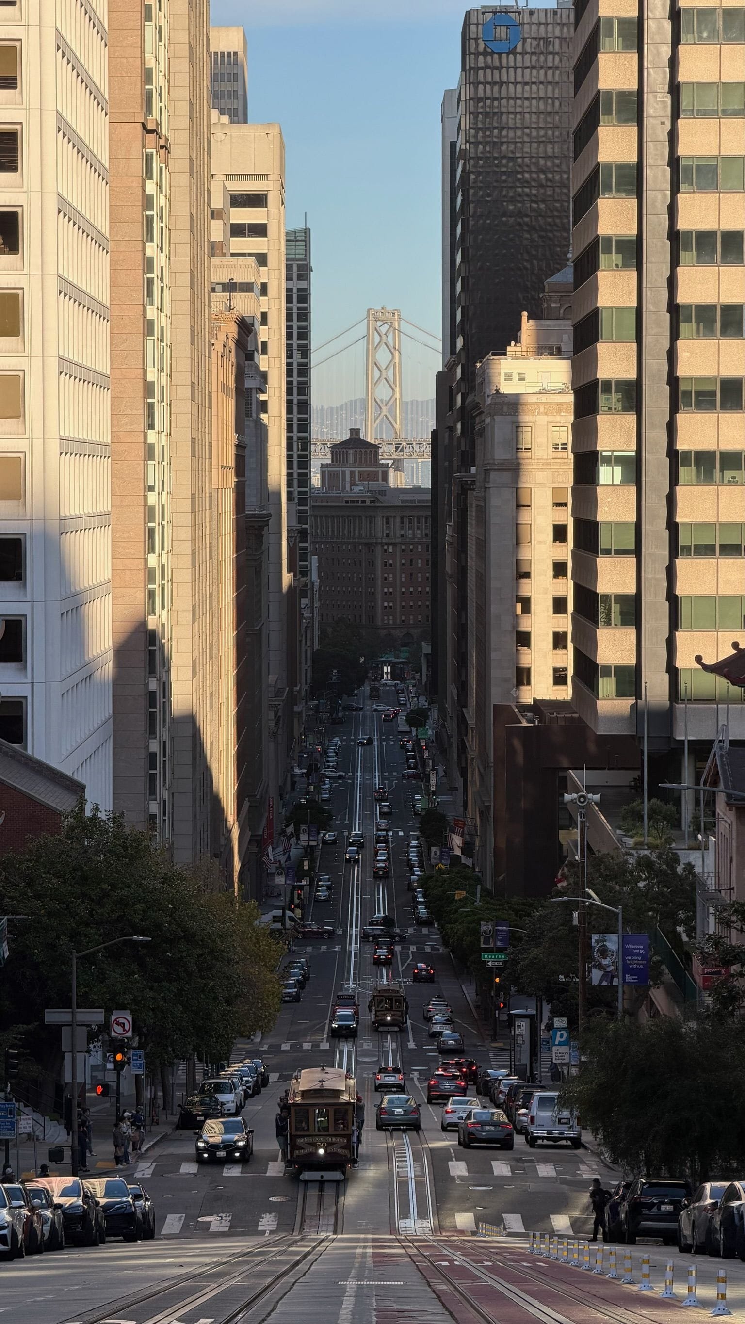 A city street view with tall buildings on both sides, filled with traffic including cars and a cable car, with the San Francisco Bay Bridge visible in the background.