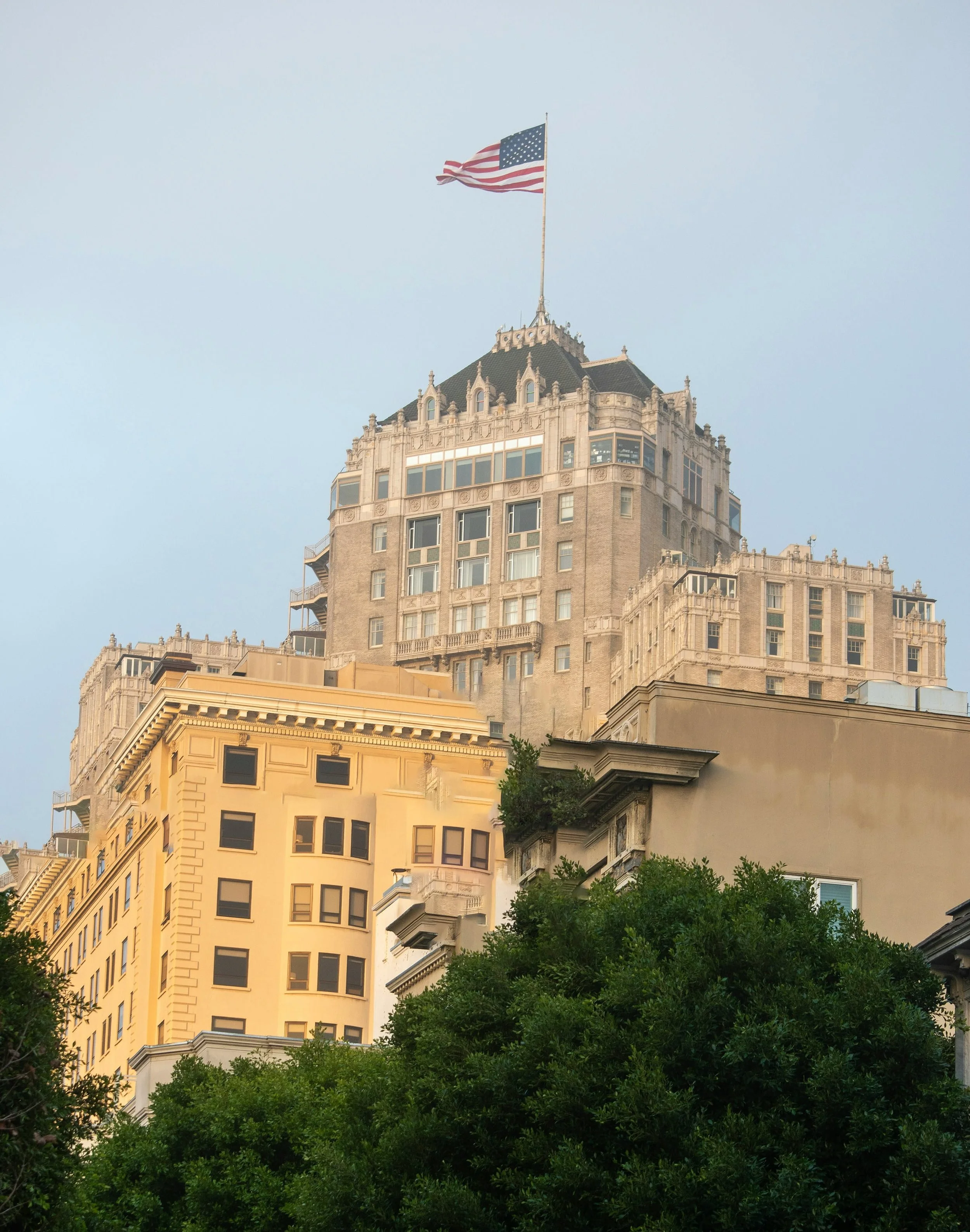 Tall historic building with a rooftop flagpole displaying the American flag, green trees at the base, and a clear blue sky background.