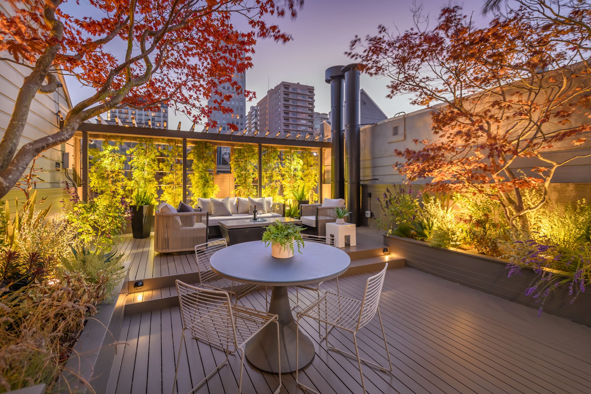 Urban rooftop patio with outdoor seating, surrounded by autumn-colored trees, illuminated wall lights, and tall city buildings in the background during dusk.