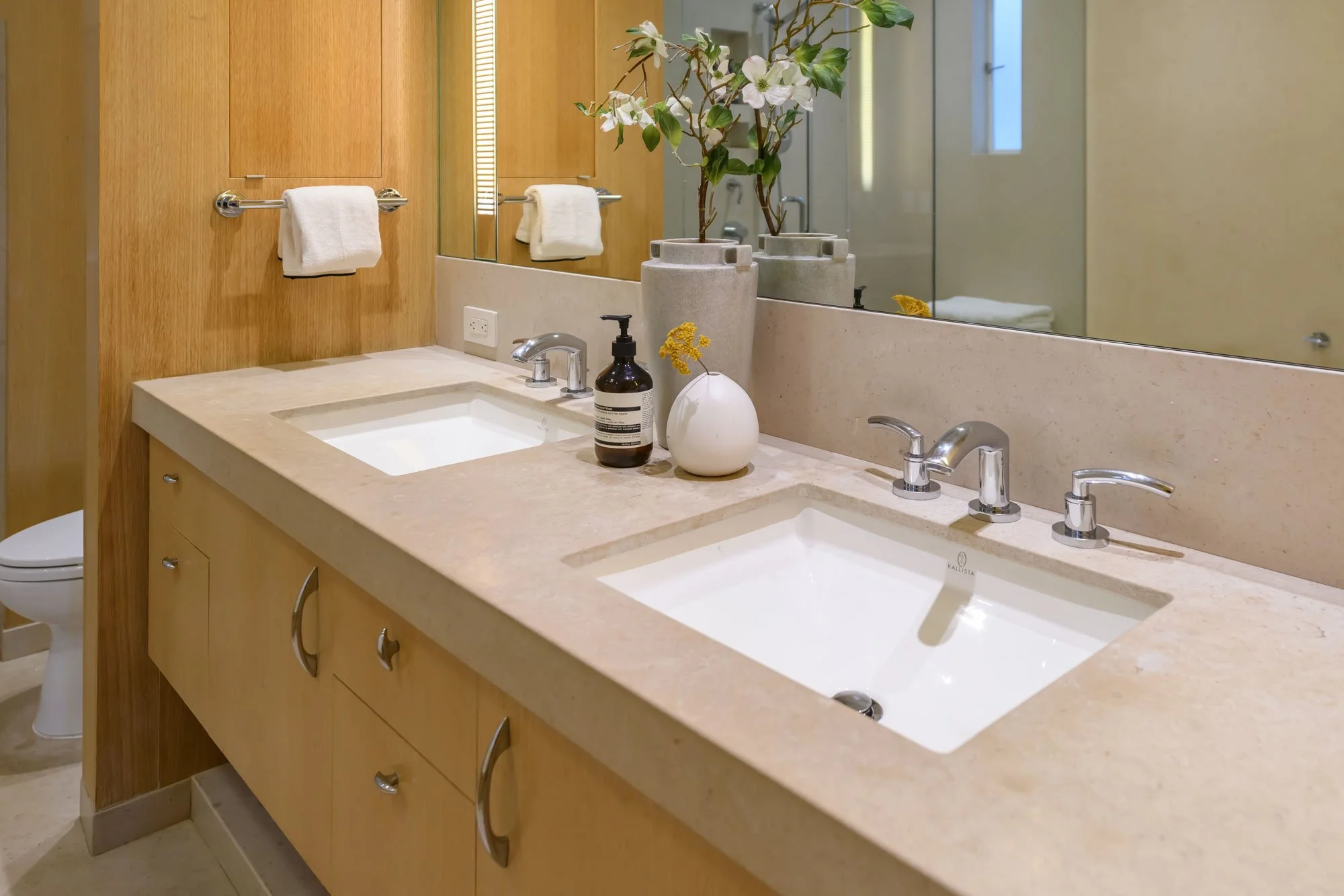 Double bathroom sink with beige countertop, large mirror, and decor including a white vase with flowers and a dark soap dispenser. Wooden cabinet below the sinks, and a towel on a chrome towel bar. Part of a toilet visible to the left.