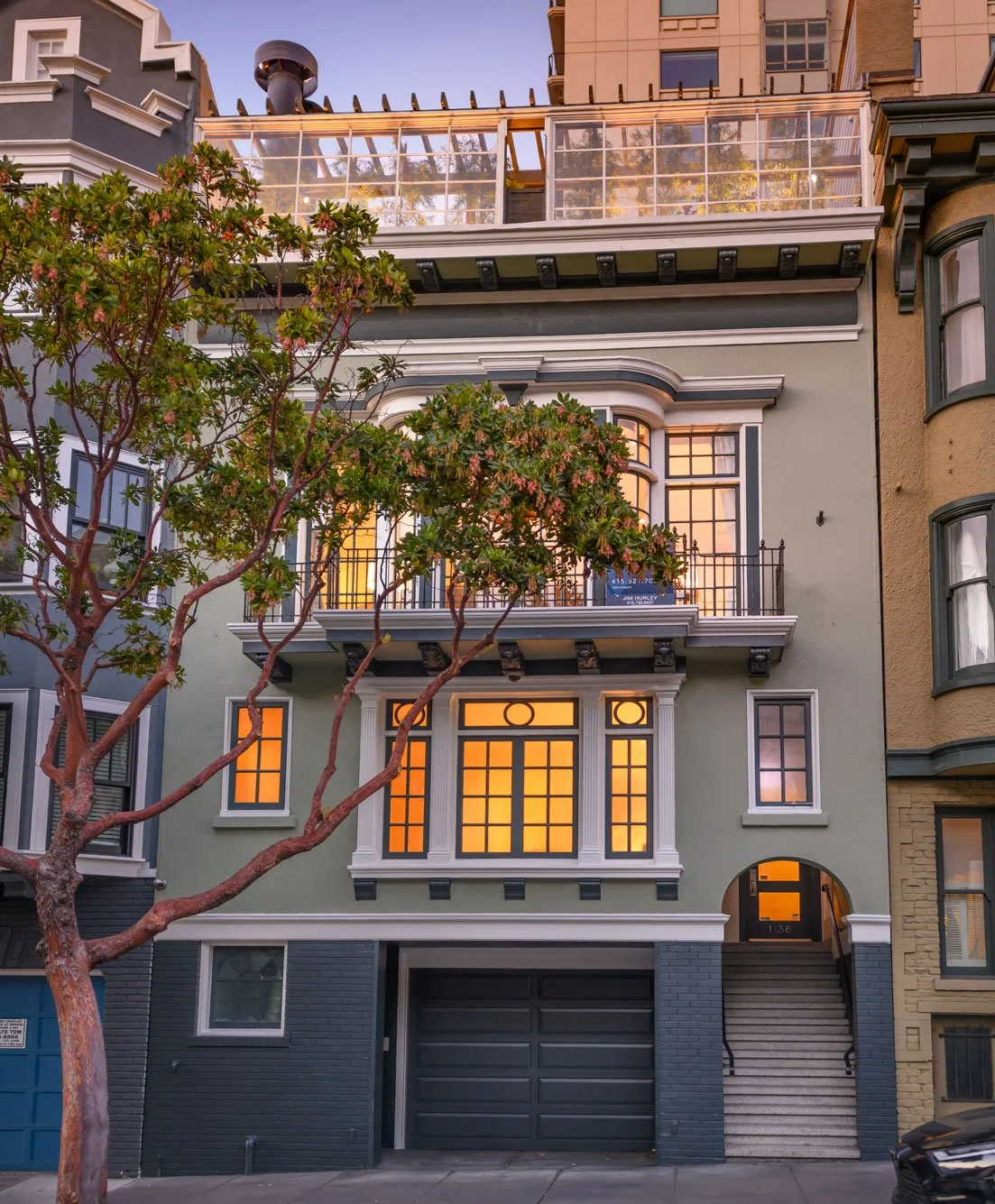 A multi-story residential building with a garage and decorative window details, lit warmly from within, with a tree in front and neighboring buildings on either side.