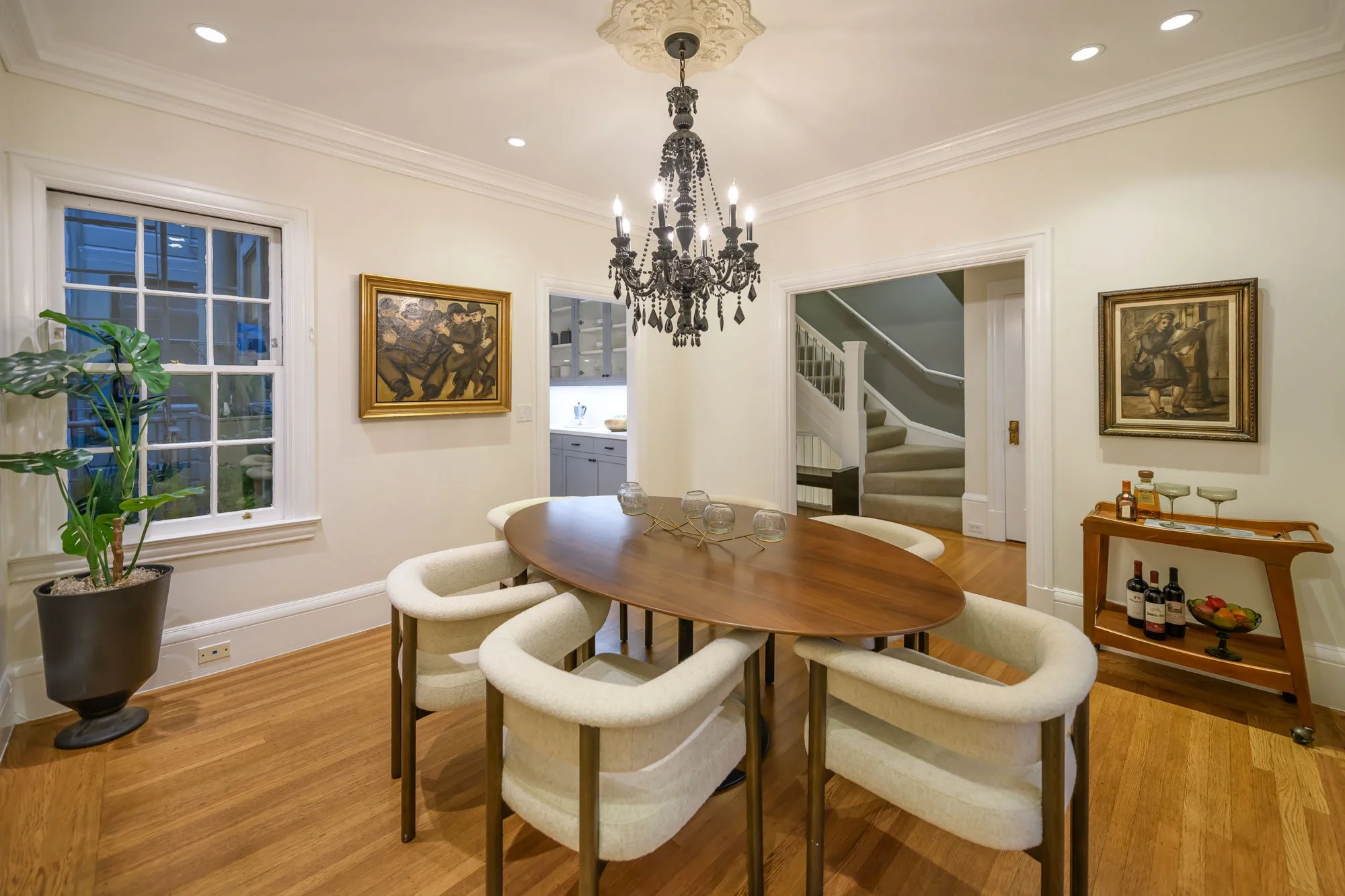 A dining room with a wooden oval table surrounded by cream-colored upholstered chairs. A black chandelier hangs from the ceiling, and the walls are decorated with framed artwork. A large window with white trim is on the left, and a small bar cart wit