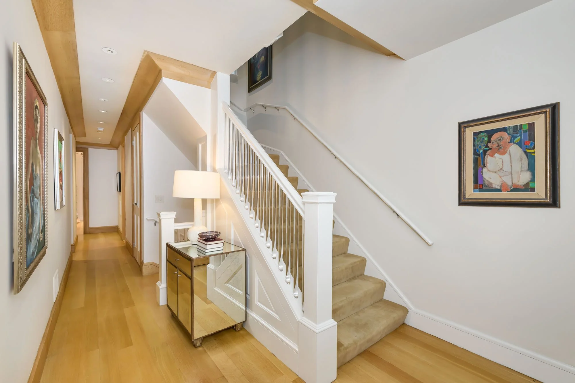 A hallway with a staircase on the right side, decorated with framed artwork on the walls, a small gold side table with a lamp and books, and wooden flooring.