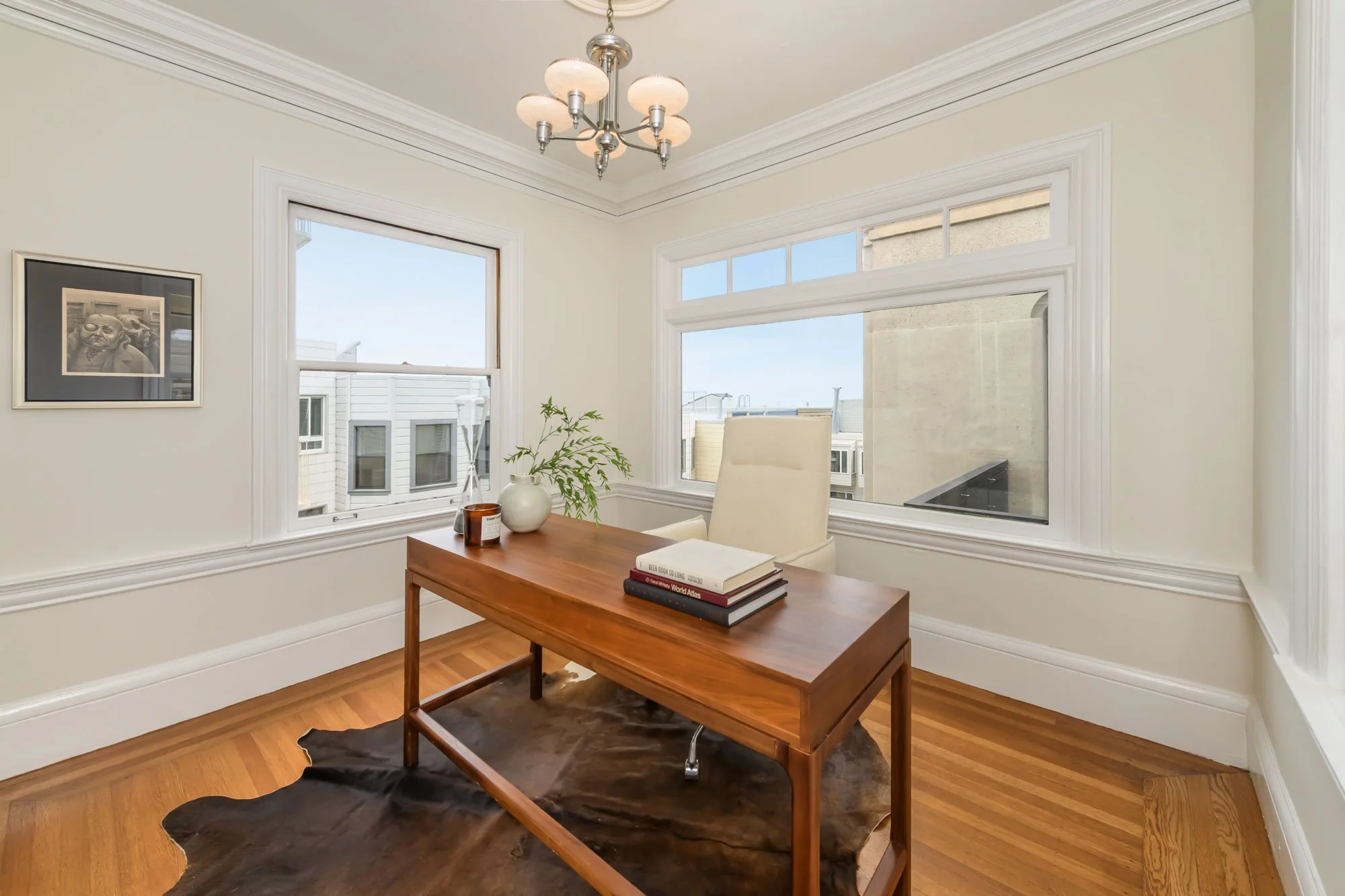 A bright dining room with a wooden table, white cushioned chairs, and large windows letting in natural light, decorated with a framed black and white photo, a white vase with greenery, and a stack of books.