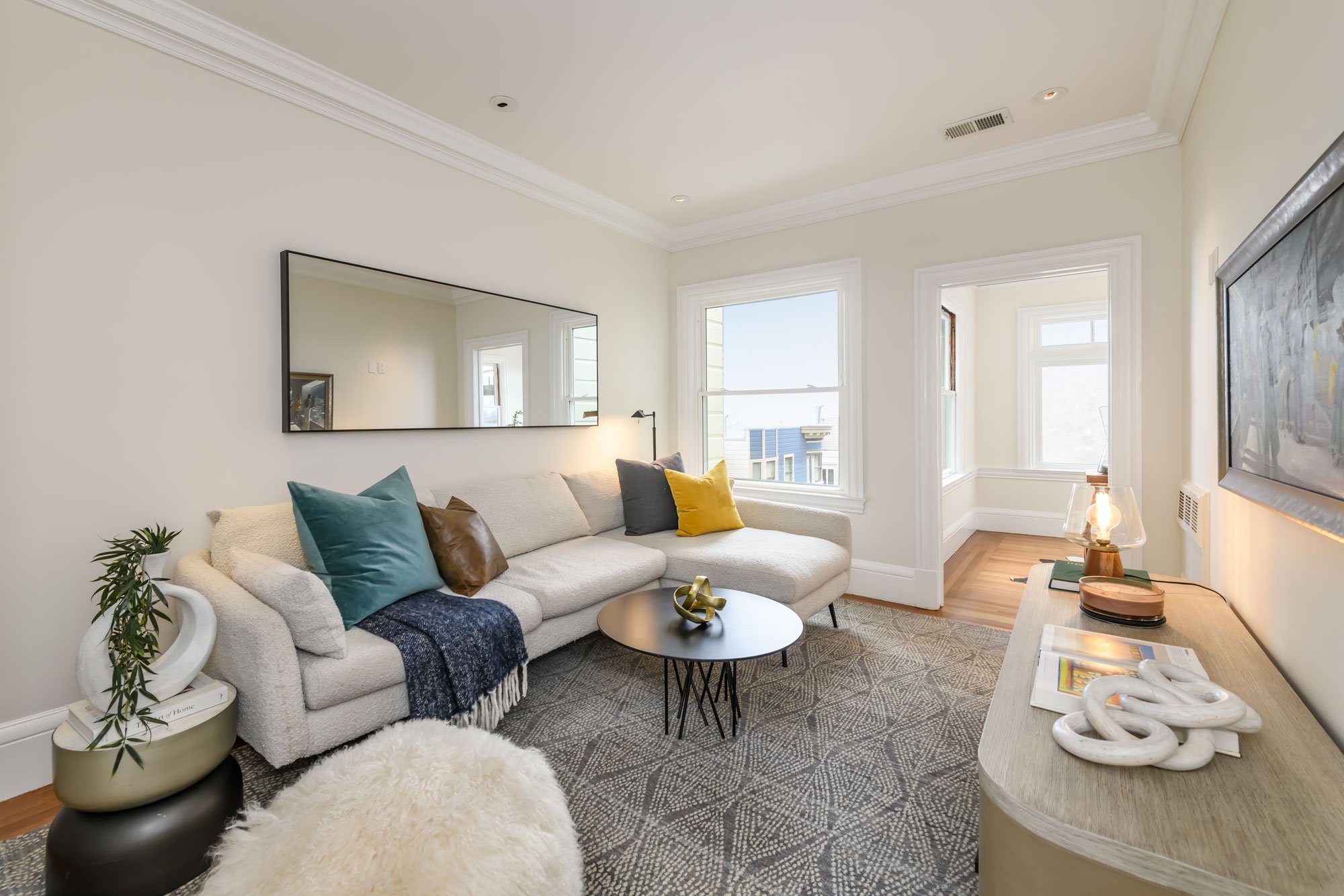 Living room with a cream sofa topped with colorful pillows, a black round coffee table, a tall plant in a beige and black pot, a large mirror on the wall, a TV, a side table with decor, and two large windows letting in natural light.