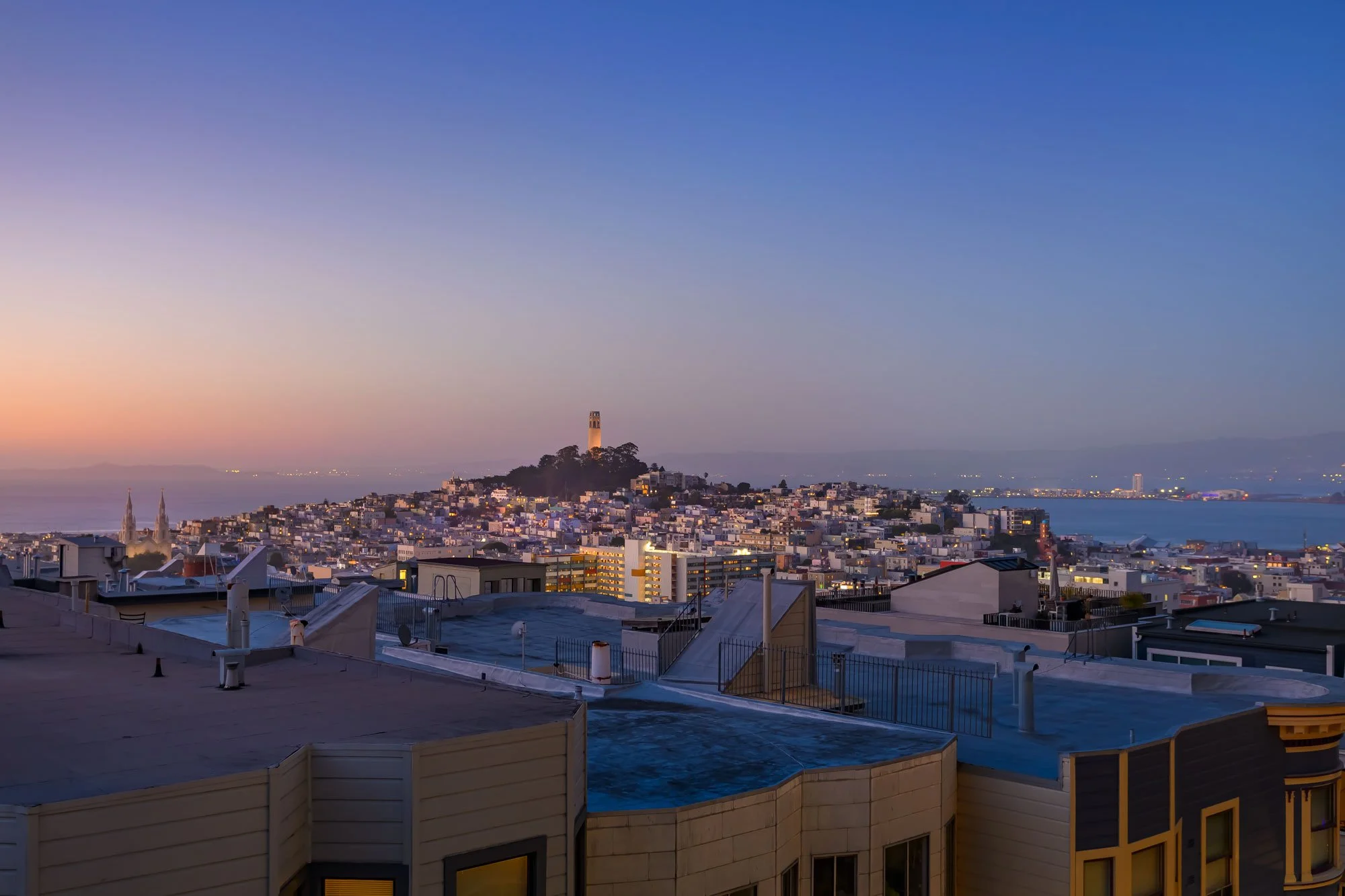 View of a city at dusk with a lighthouse on a hill in the background, modern buildings in the foreground, and a calm body of water in the distance.