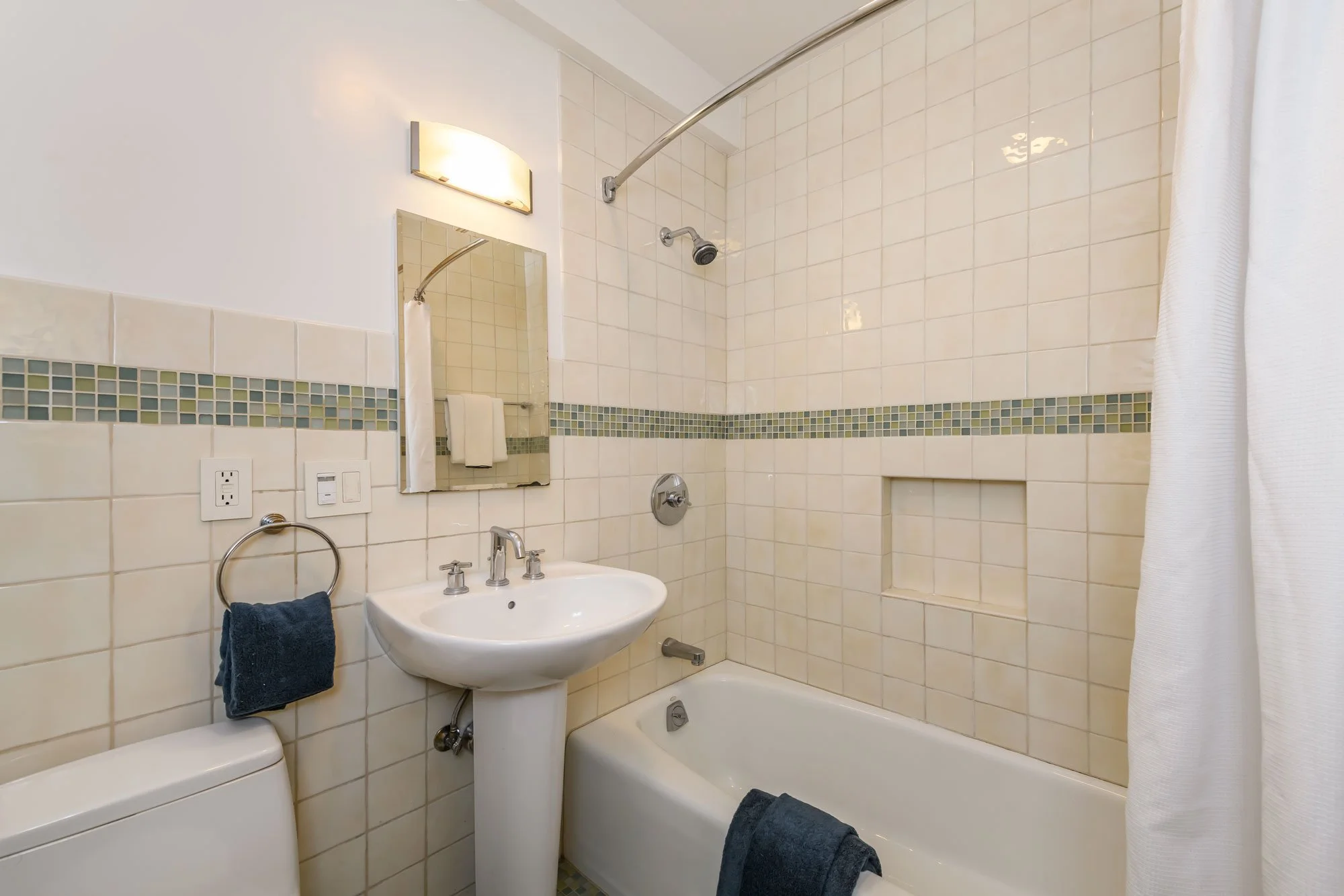 A compact bathroom with a white sink, mirror, and a small bathtub. Tiled walls with a decorative border, a towel hanging on a ring, and a light fixture above the mirror. Including a toilet and a showerhead.