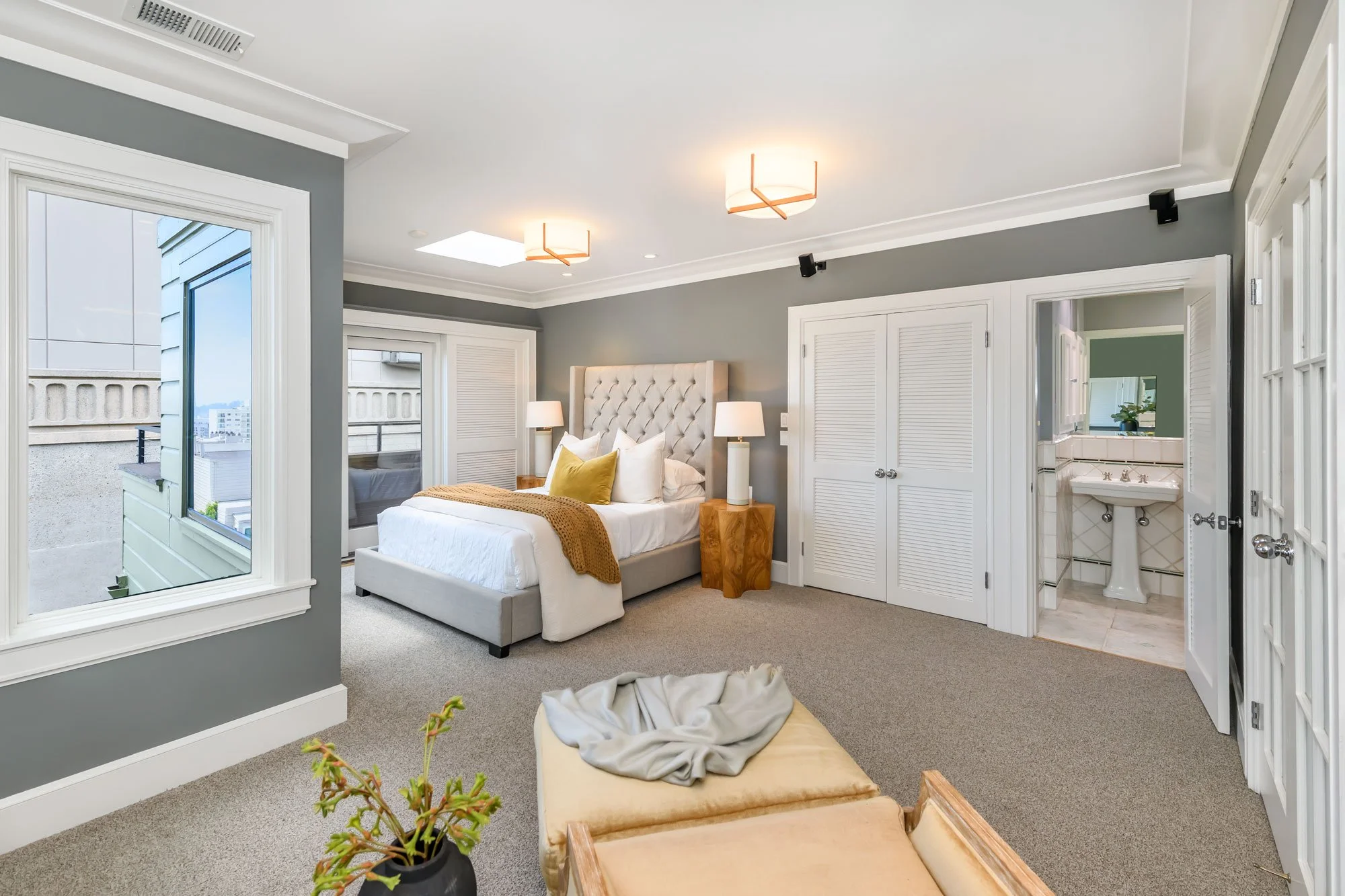 A modern bedroom with a light-colored upholstered bed, two bedside lamps, and a window showing a city view. An open door reveals an adjoining bathroom with a pedestal sink and mirror. Neutral gray walls with white trim, ceiling lights, and a small po