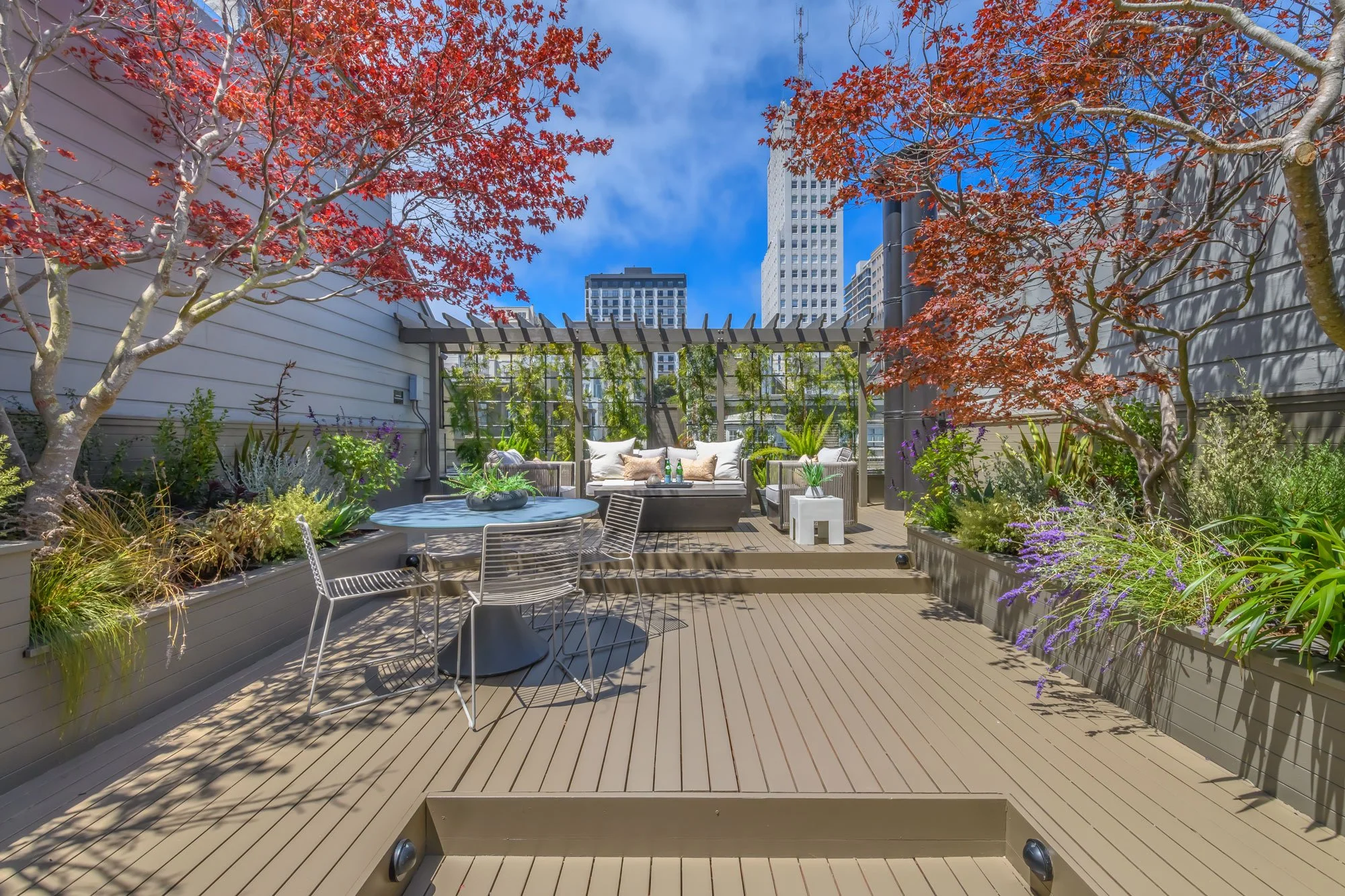 Urban rooftop terrace with deck flooring, surrounded by trees with red and purple leaves, metal patio furniture, a white cushioned seating area, and a city skyline in the background under a blue sky.