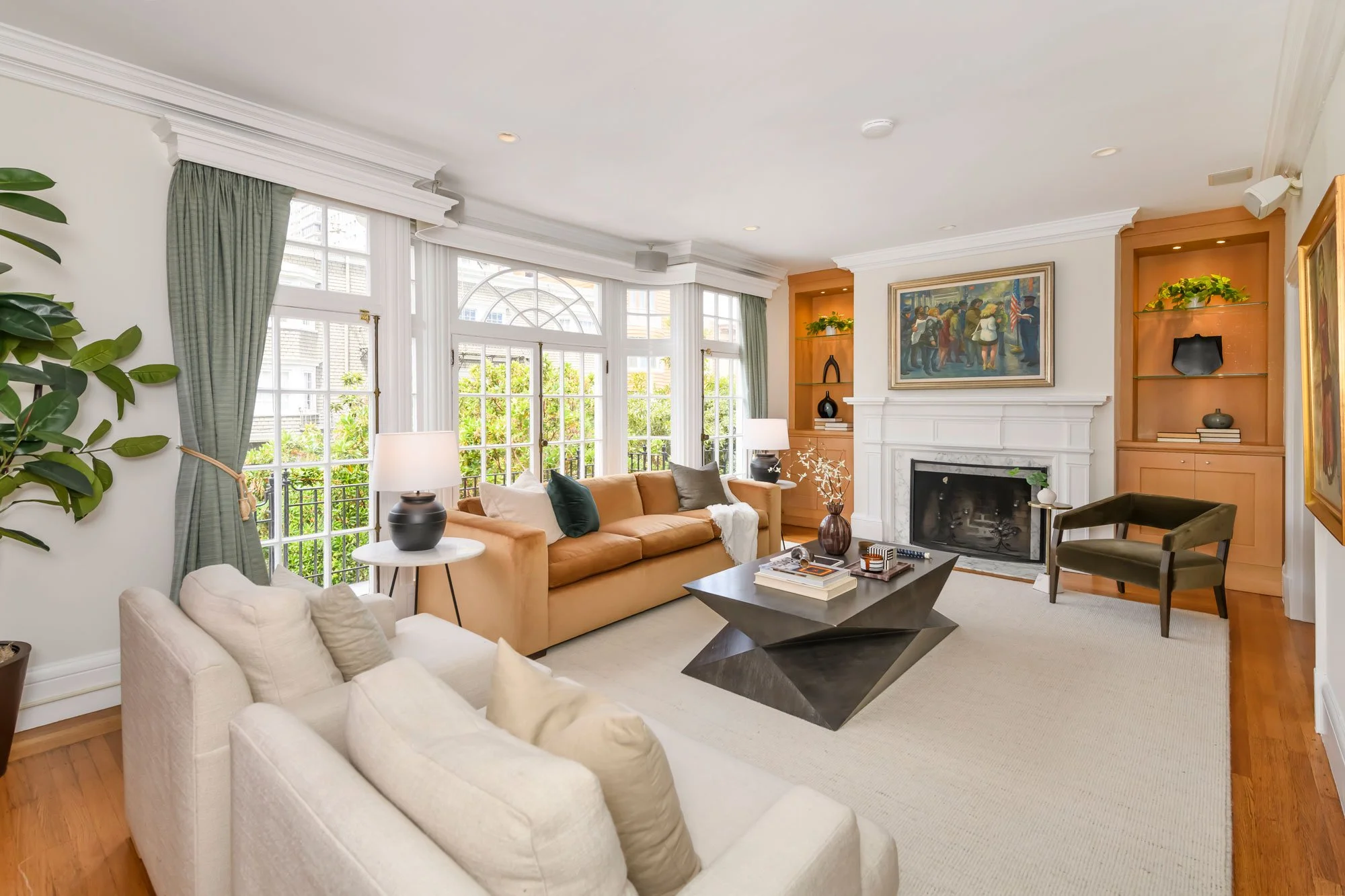 Living room with beige sofas, black coffee table, large windows with green curtains, fireplace, wall art, built-in shelves with decorations, potted plant, and hardwood flooring.