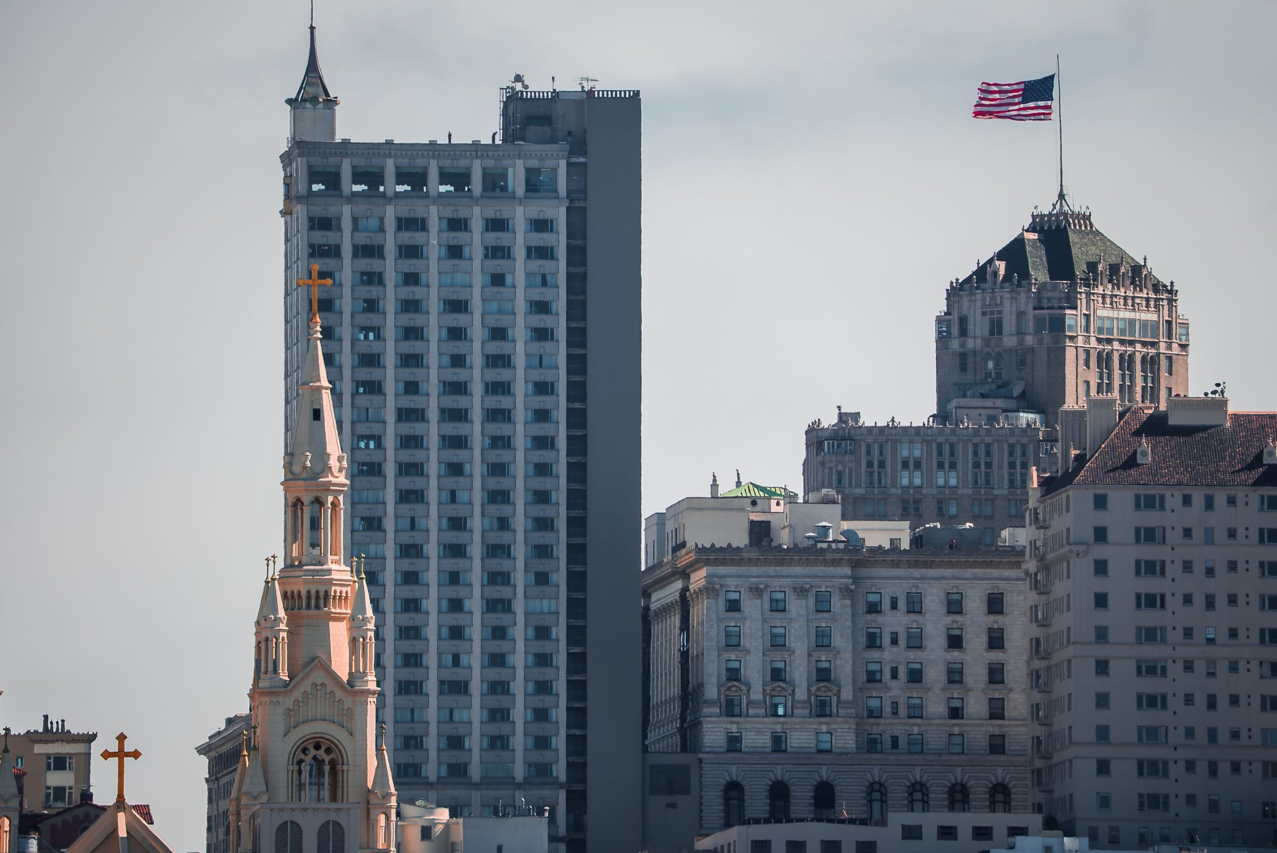 City skyline featuring historic church with light pink facade and green roof, tall modern skyscrapers, and an American flag flying on a flagpole.