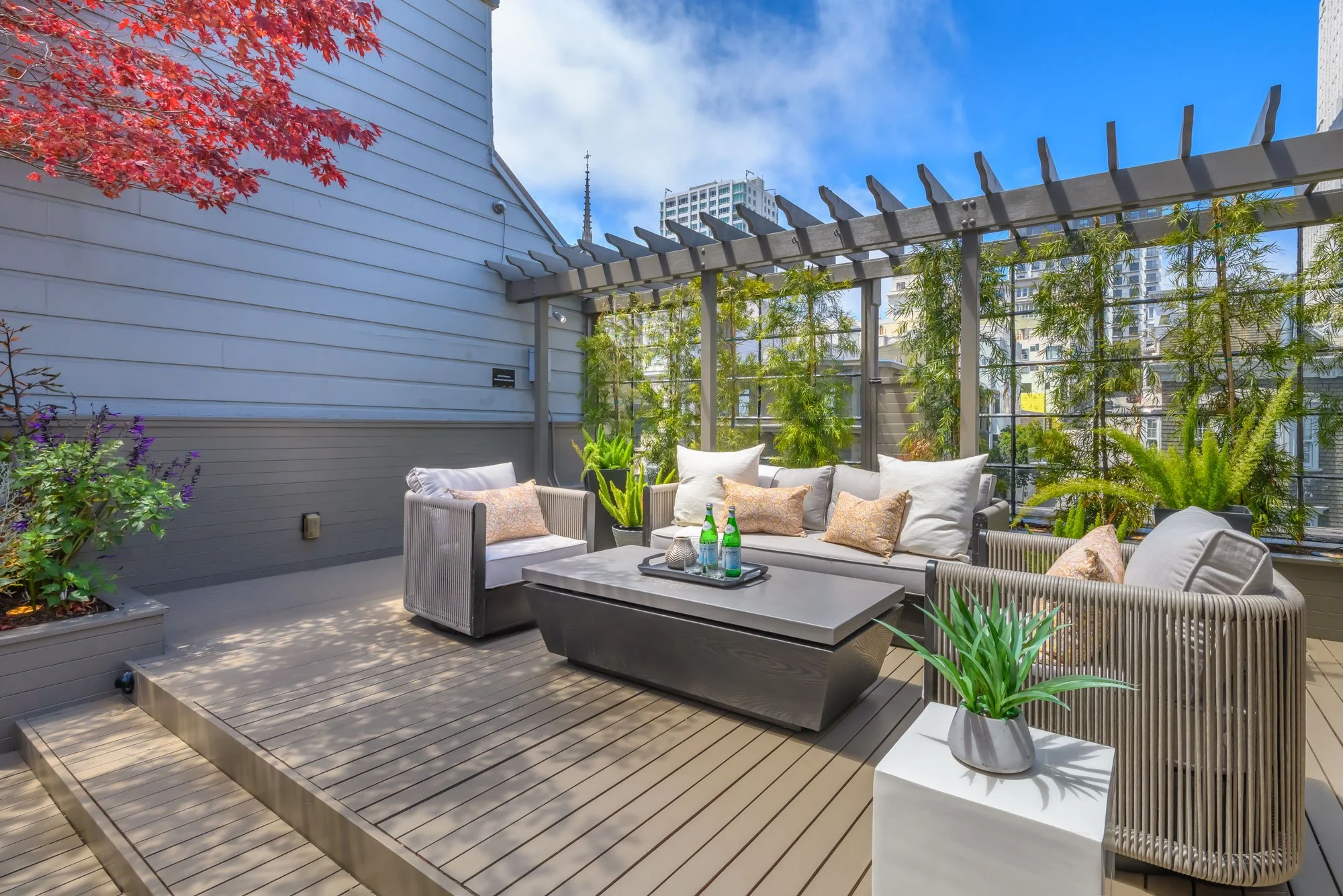 Urban rooftop patio with beige outdoor furniture, potted plants, and greenery, overlooking city buildings under a blue sky.