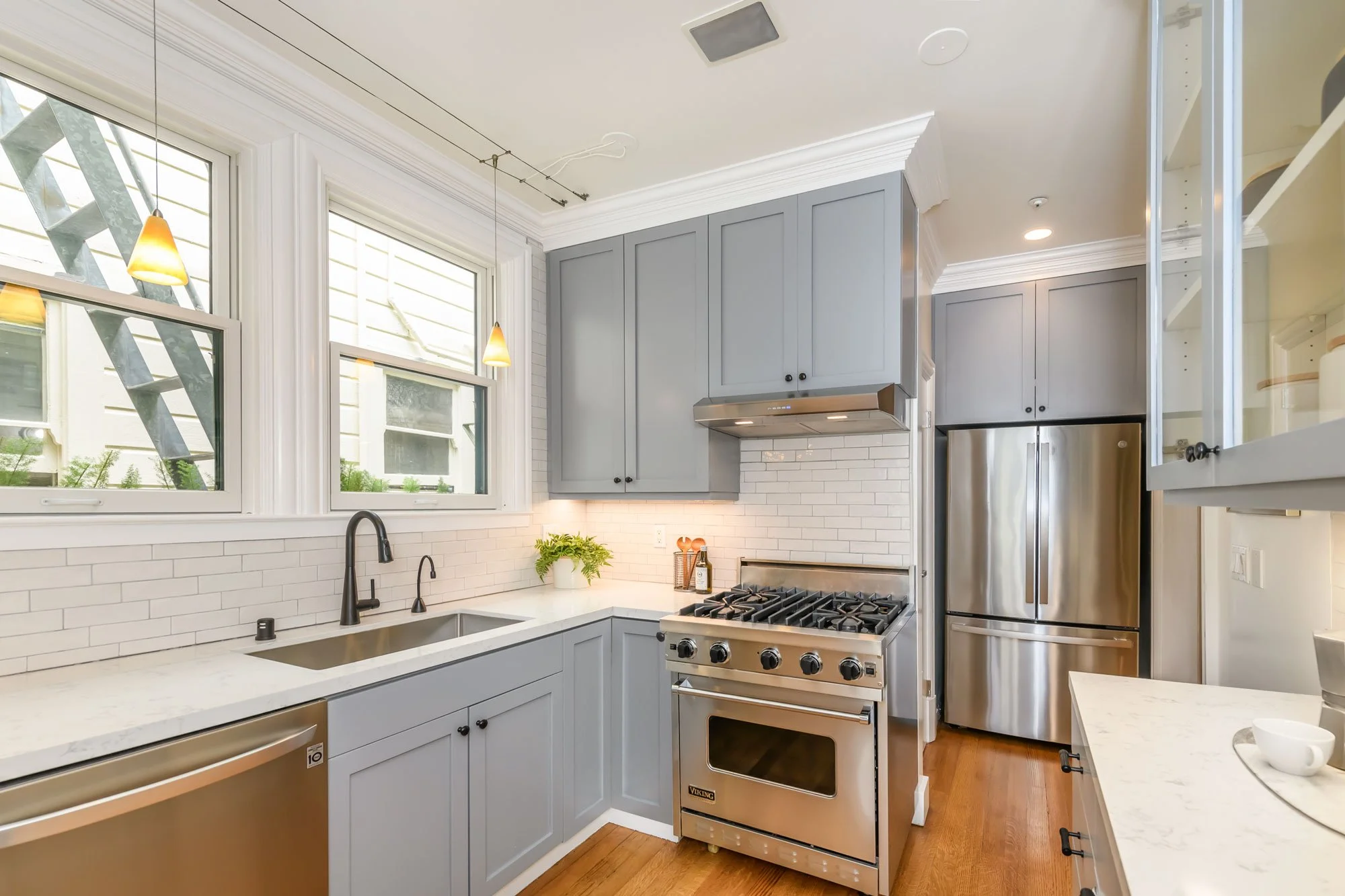 Modern kitchen with gray cabinetry, white subway tile backsplash, stainless steel appliances including a refrigerator and oven, black faucet, and two pendant lights with warm yellow shades.
