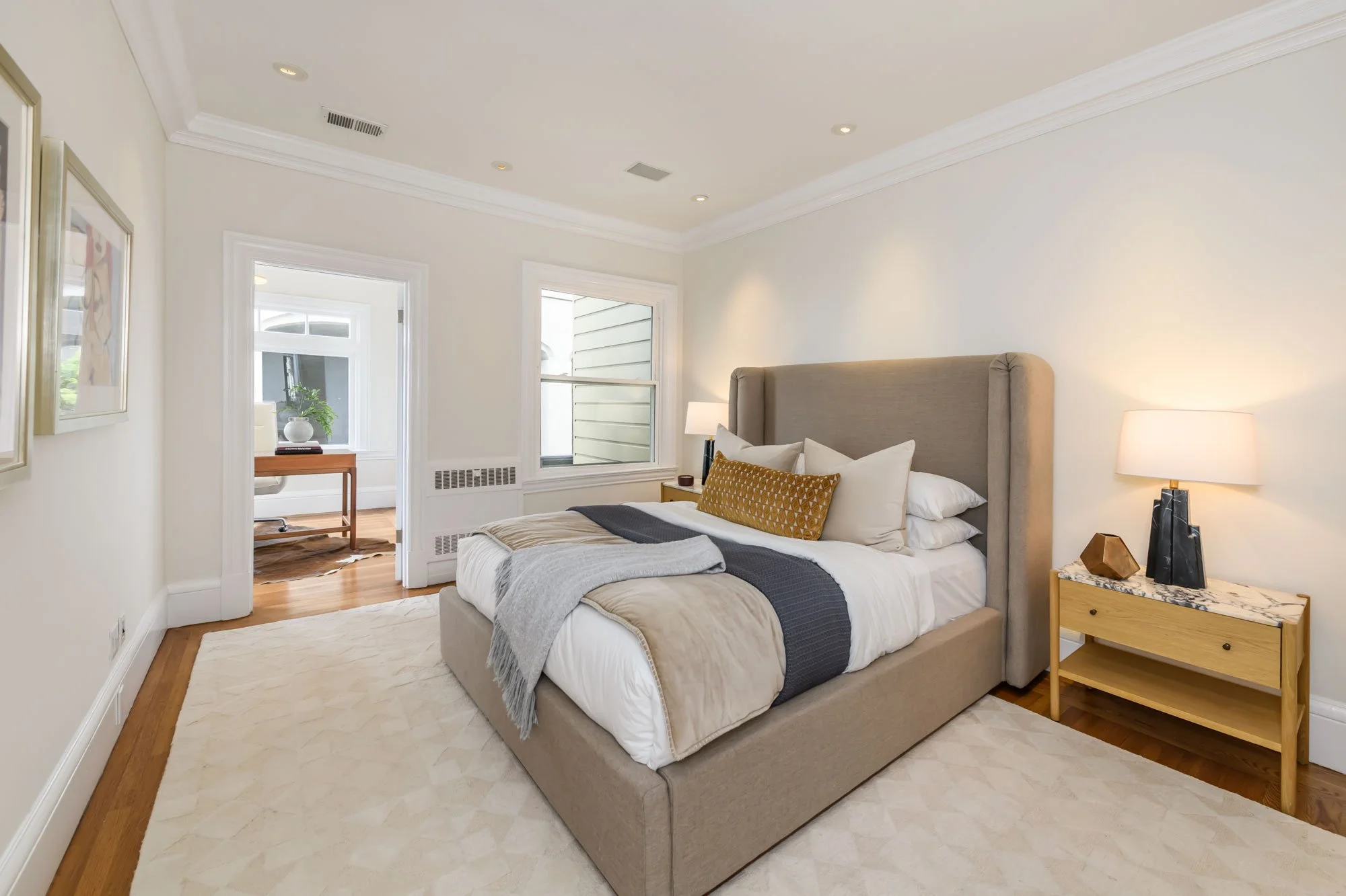 Modern bedroom with beige upholstered bed, white bedding, decorative pillows, nightstand with lamps and decor, hardwood floors, white walls and crown molding, and a window with sunlight.