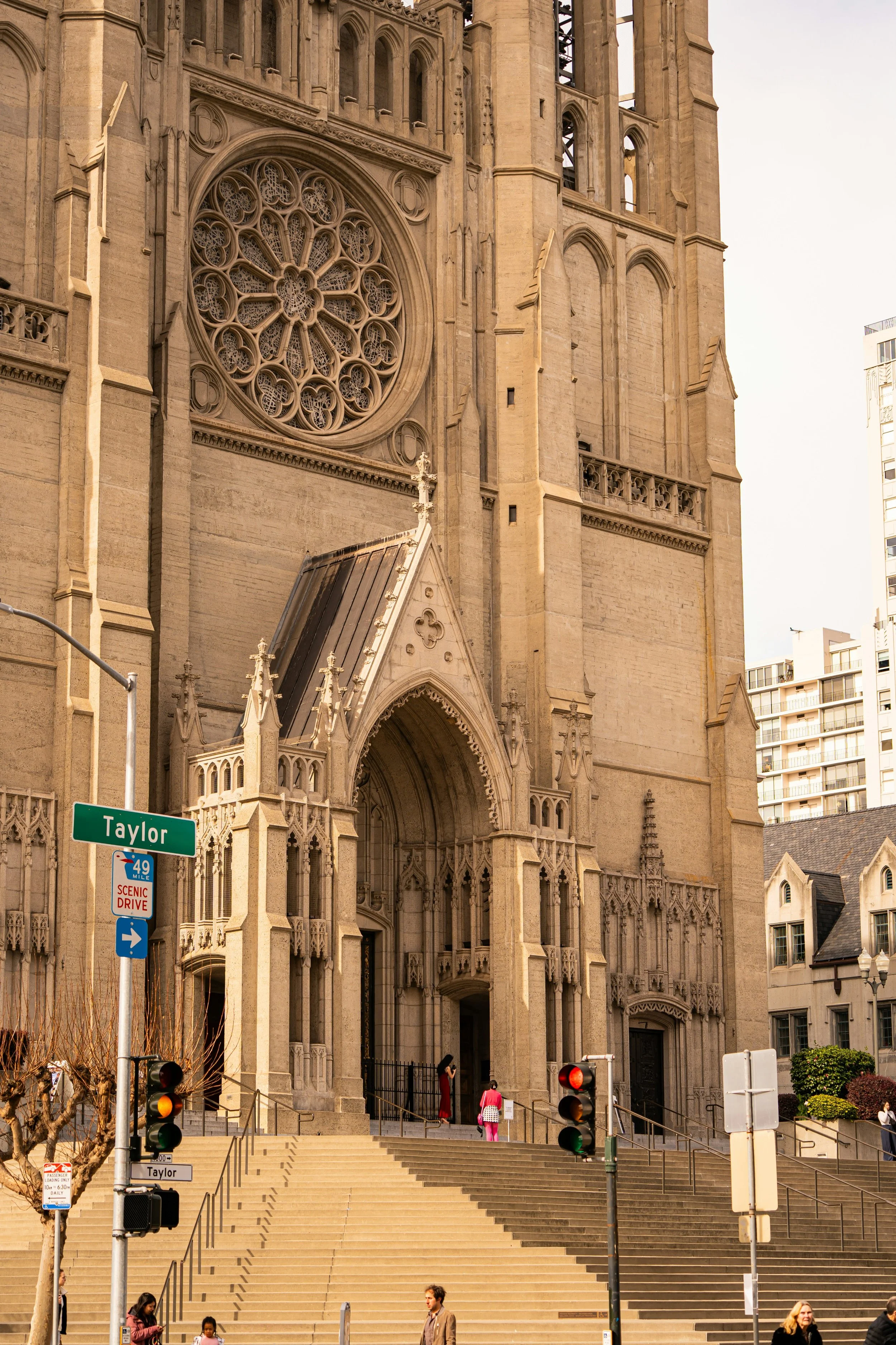 Front of a large historic church in a city, with steps leading up to the entrance, traffic lights, and street signs including one that says 'Taylor' and a scenic drive sign. Several people are walking near the stairs.