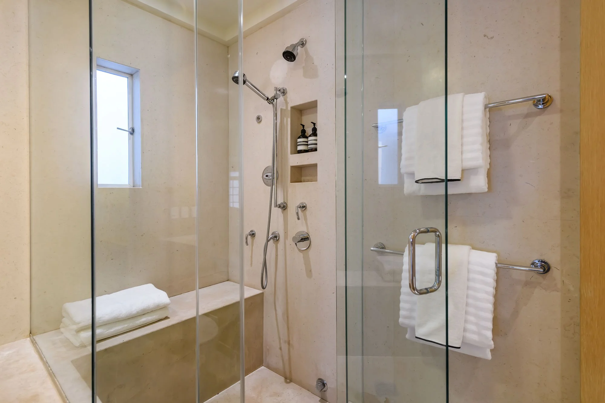 A modern glass-enclosed shower with beige marble walls and floor, chrome showerheads, and a built-in niche holding two bottles of soap or shampoo. There are neatly folded white towels on a wooden bench outside the shower. A small frosted window provi
