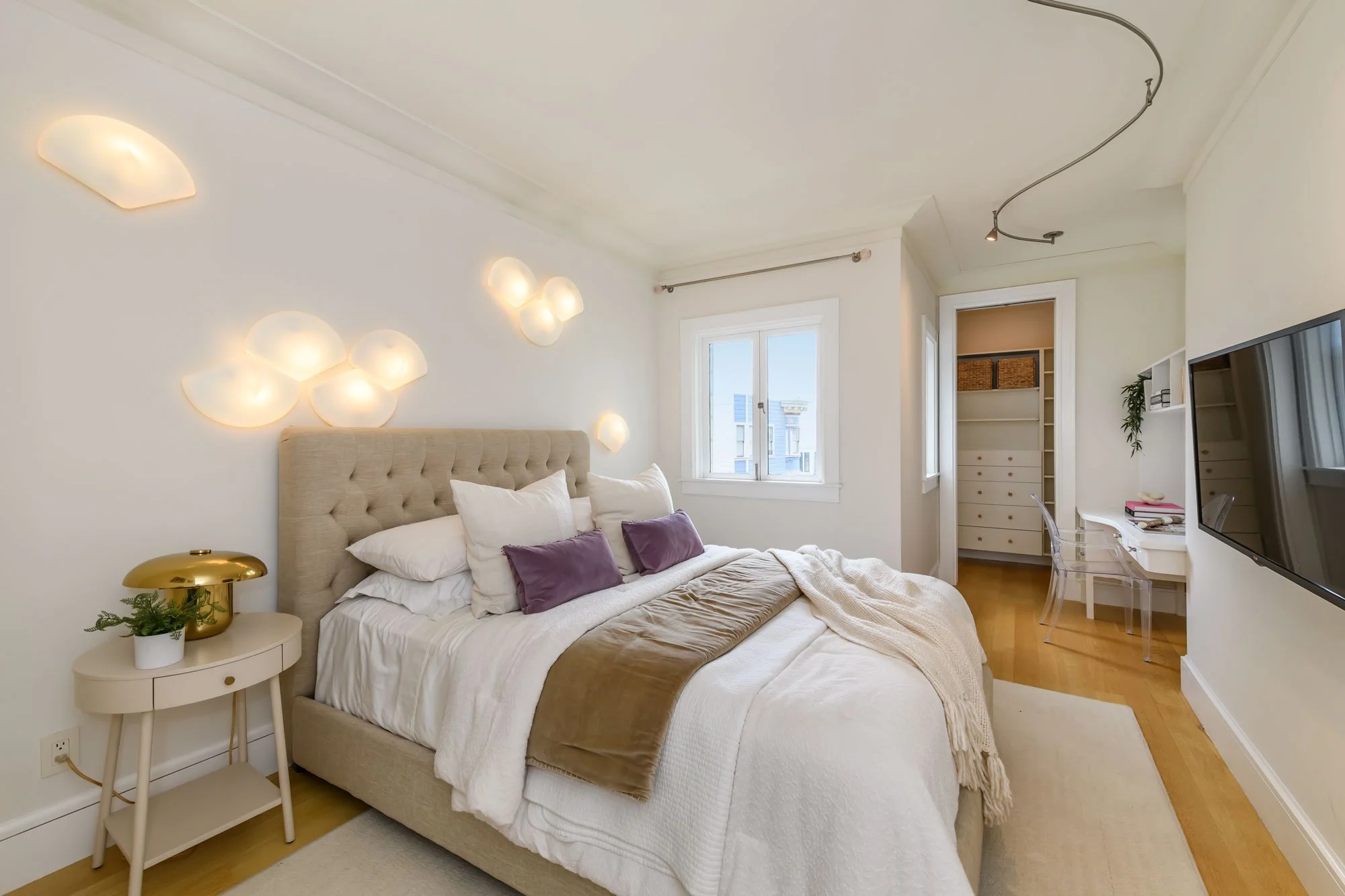 A bedroom with a bed adorned with white and purple pillows, a beige headboard, and a white nightstand with a gold lamp and small plant. There are modern wall sconces above the bed, a window with a view of neighboring buildings, and a closet with open