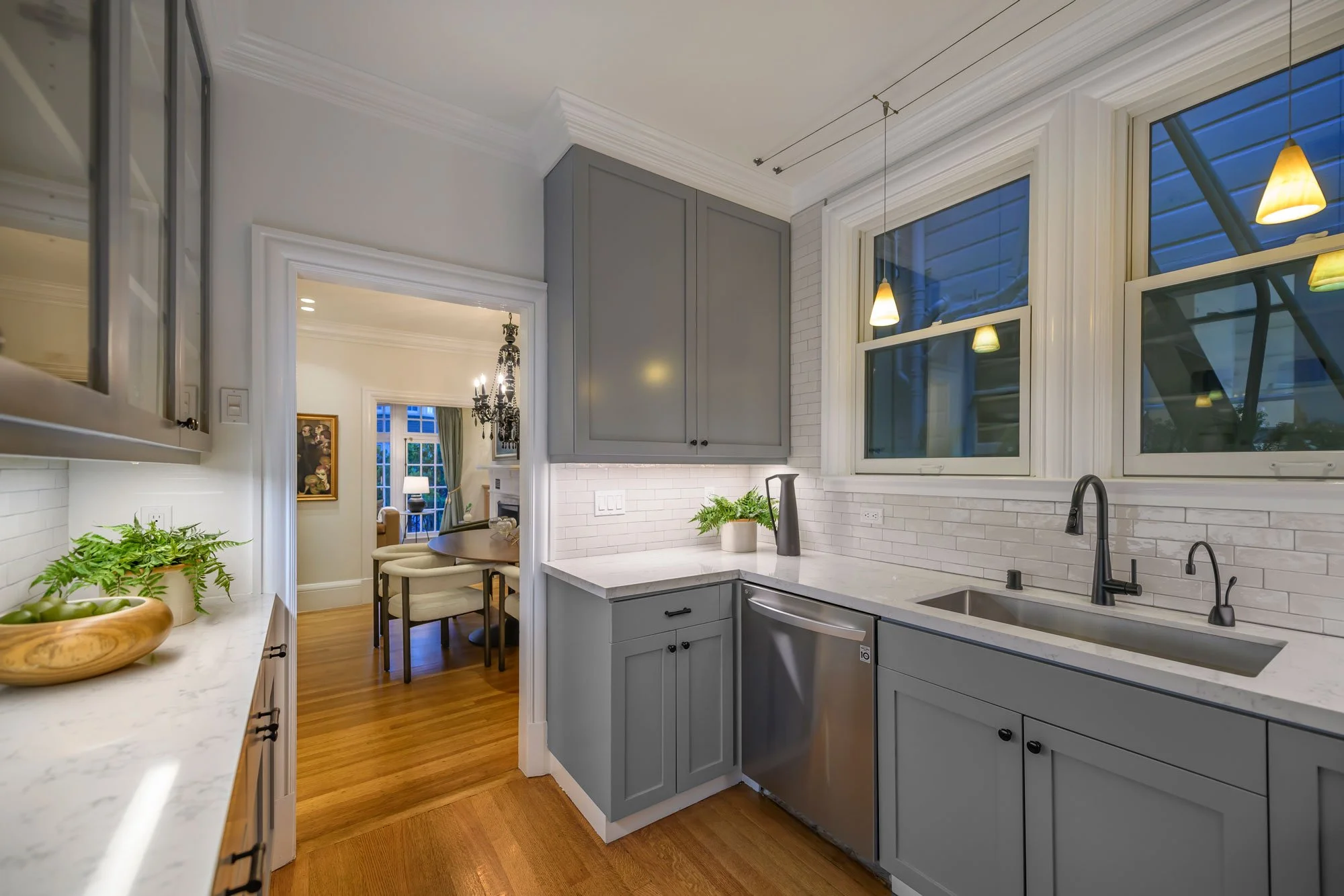 Kitchen with gray cabinets, white marble countertops, large windows, and pendant lights. Open doorway leads to a dining area with a chandelier and a painting.