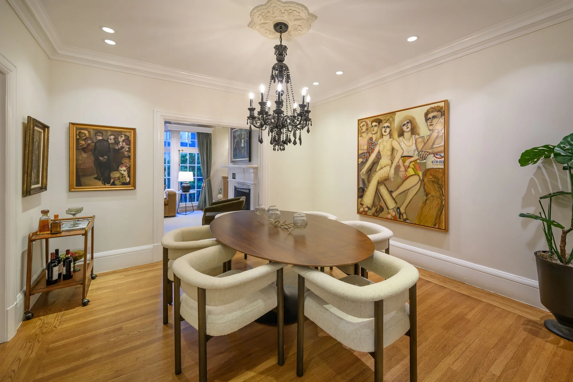 A dining room with an oval wooden table surrounded by cream-colored upholstered chairs, a black chandelier hanging from a decorative ceiling medallion, paintings on the cream walls, and a window with green curtains in the background.