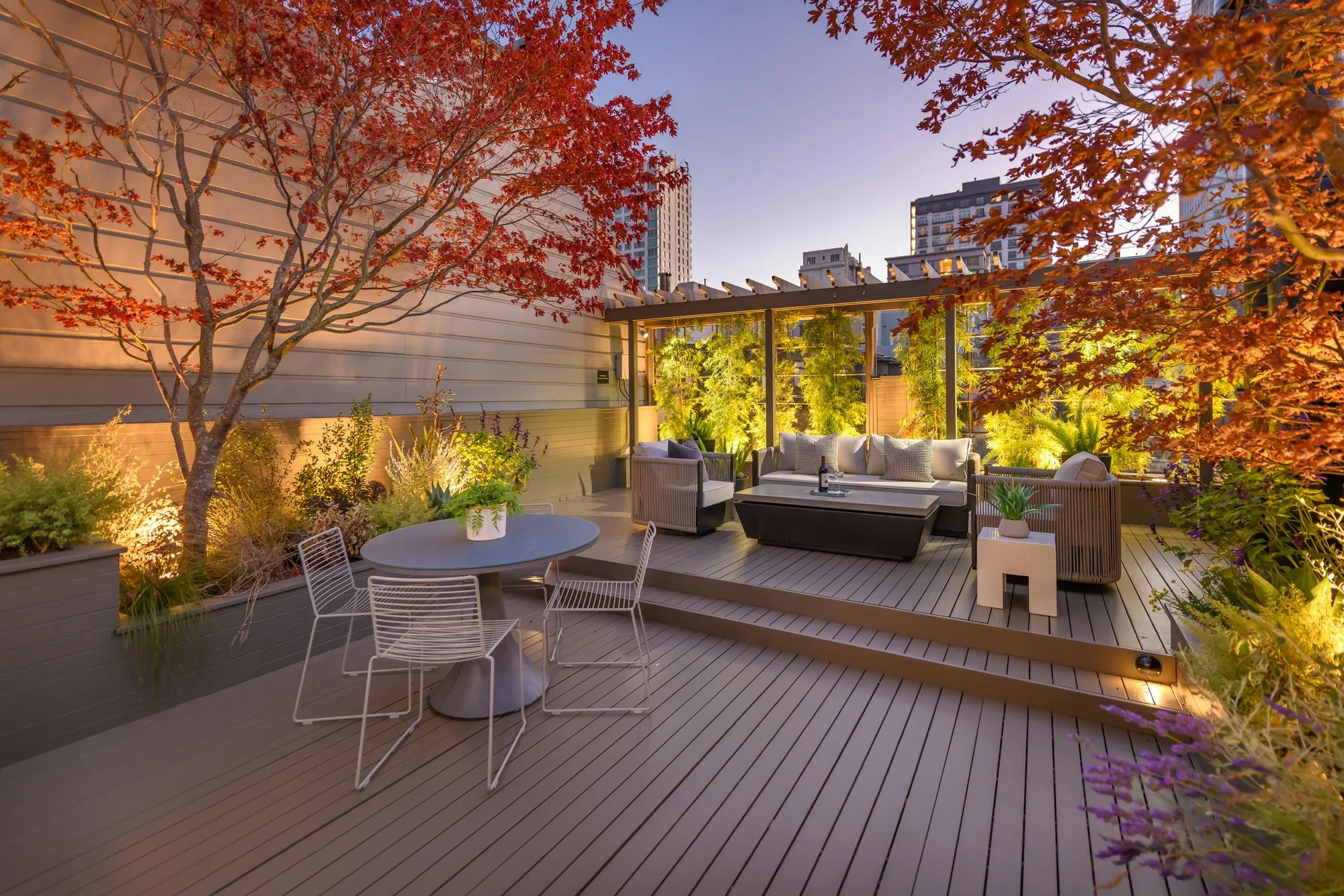 A rooftop patio with a large red maple tree, modern furniture, potted plants, and city buildings in the background during dusk.