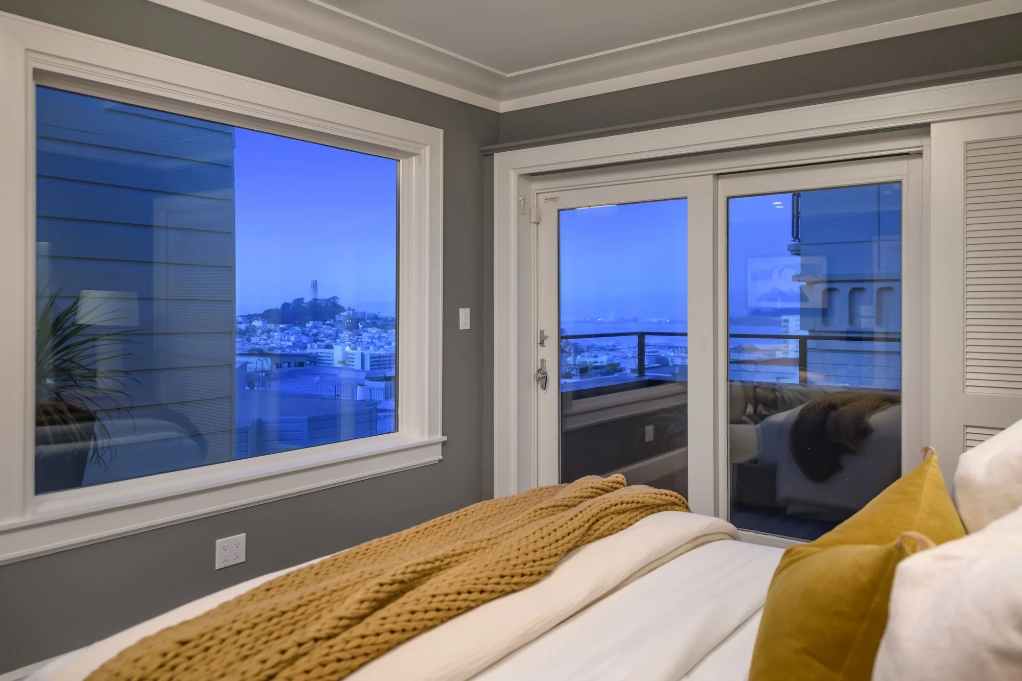 A bedroom with large windows showing a cityscape view, including a hill with a tower, and a balcony. The bed has a beige knitted throw and a yellow cushion.