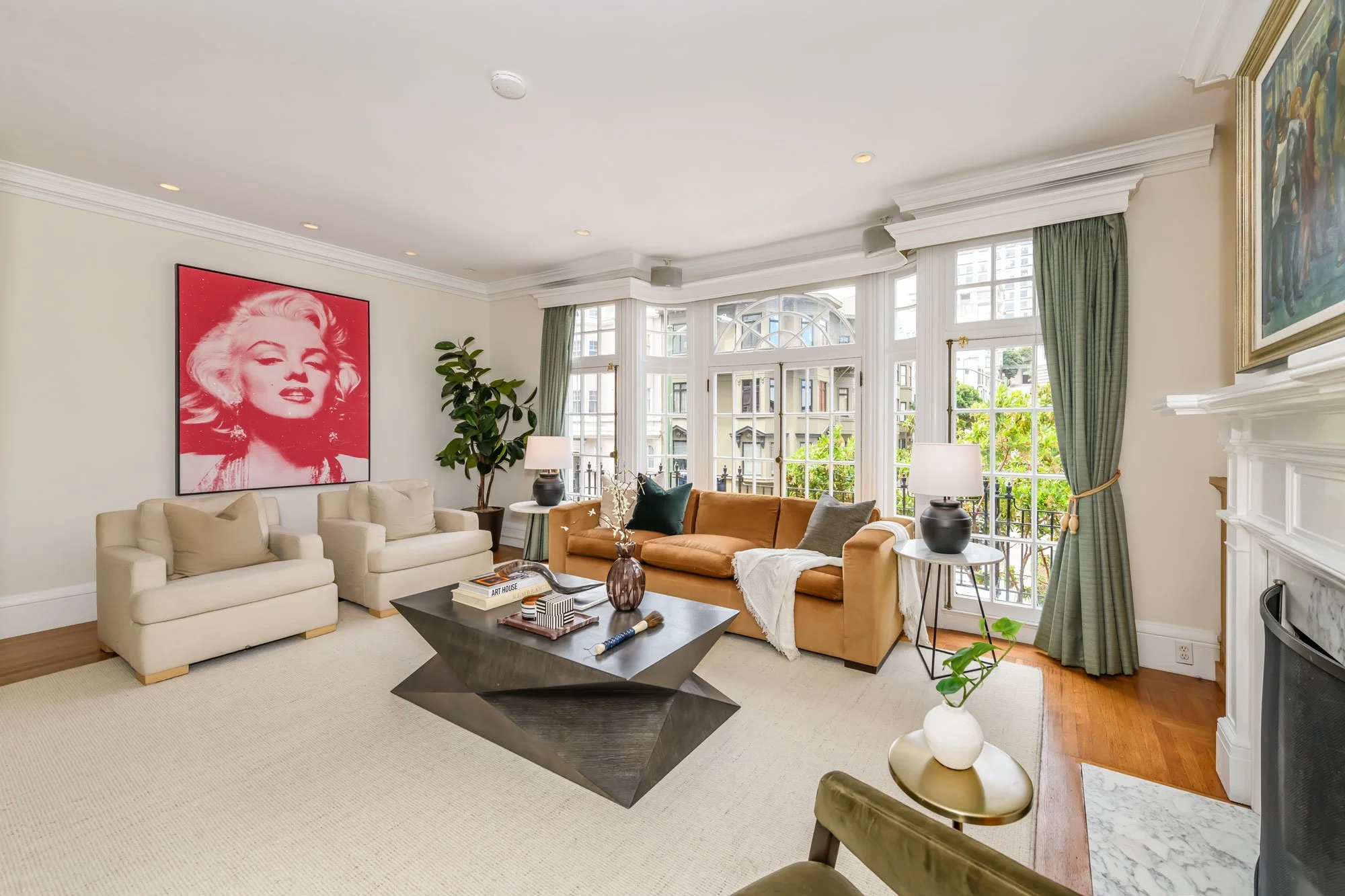 Living room with beige and orange sofas, coffee table, large windows with green curtains, wall art of Marilyn Monroe, potted plant, lamps, and fireplace.