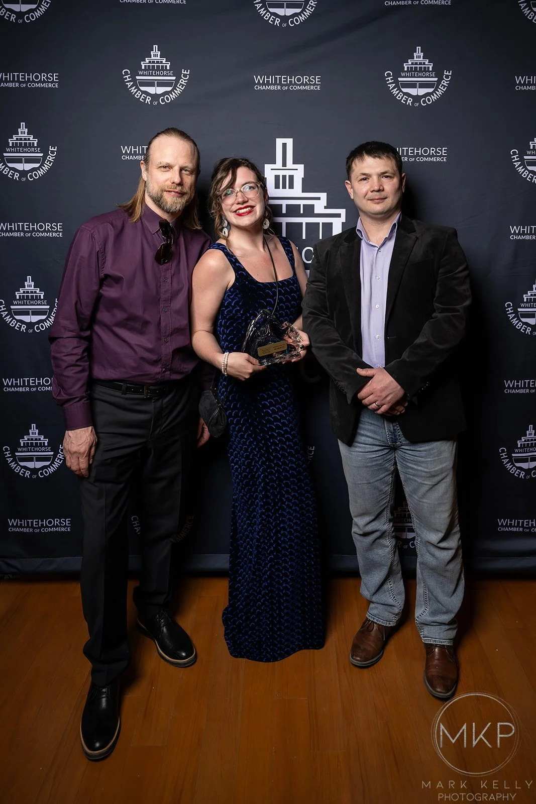 Three people stand in front of a backdrop with the Whitehorse Chamber of Commerce logo. The woman in the middle is holding a glass award and is wearing a dark blue dress, flanked by two men in business casual attire.