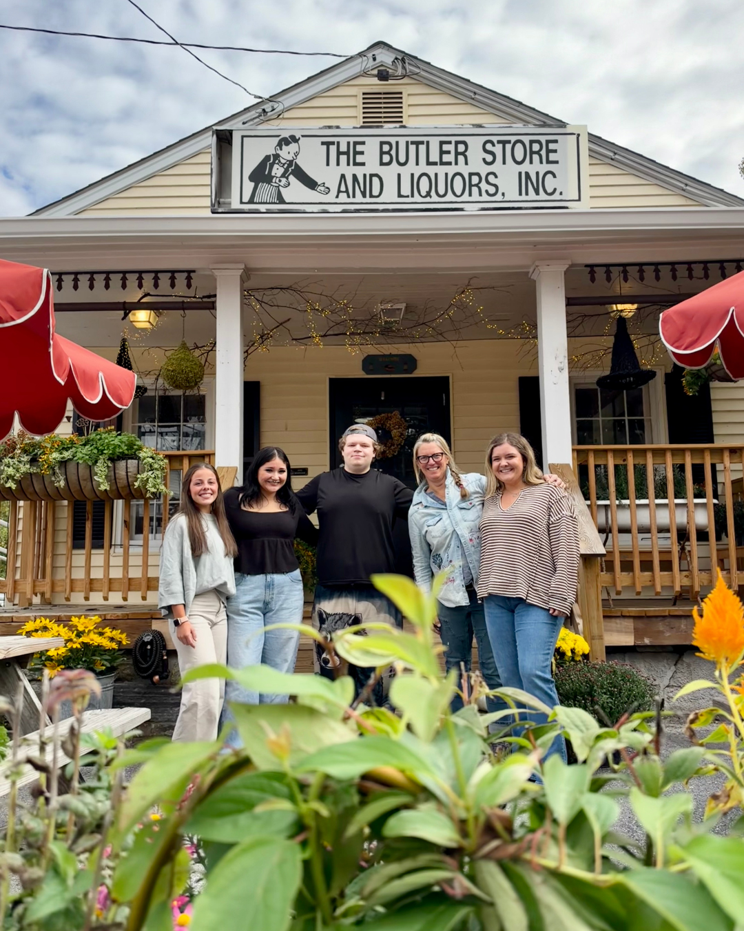 A group of five people standing on a porch in front of The Butler Farm Store, with plants and flowers in the foreground and a cloudy sky overhead.