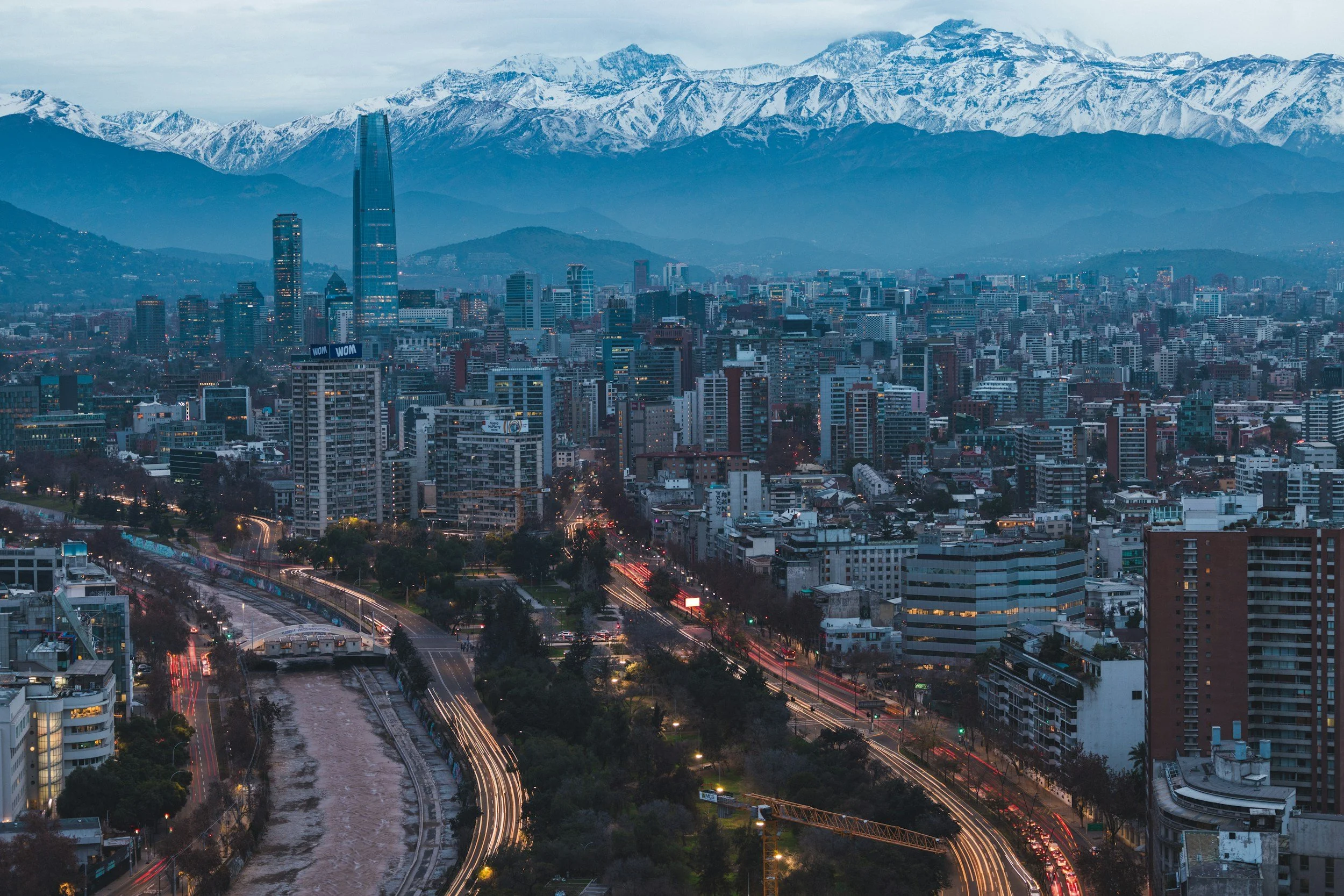 A cityscape with tall buildings and skyscrapers, mountains with snow in the background, a river, and streets with moving cars.