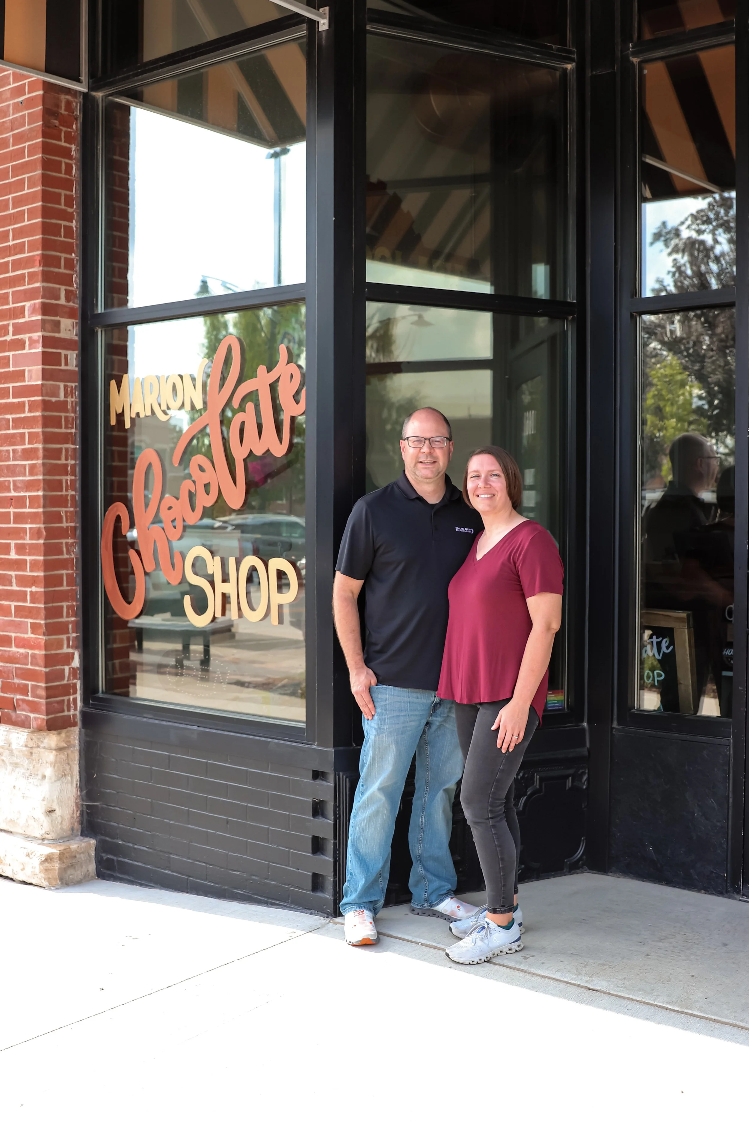 A man and a woman standing outside a chocolate shop, smiling at the camera