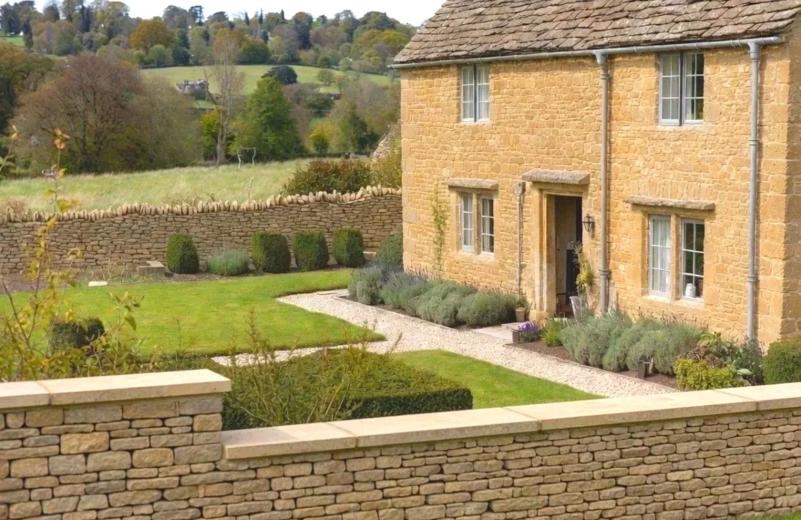 A stone house with a garden and a stone wall, surrounded by rolling green hills and trees in the background.