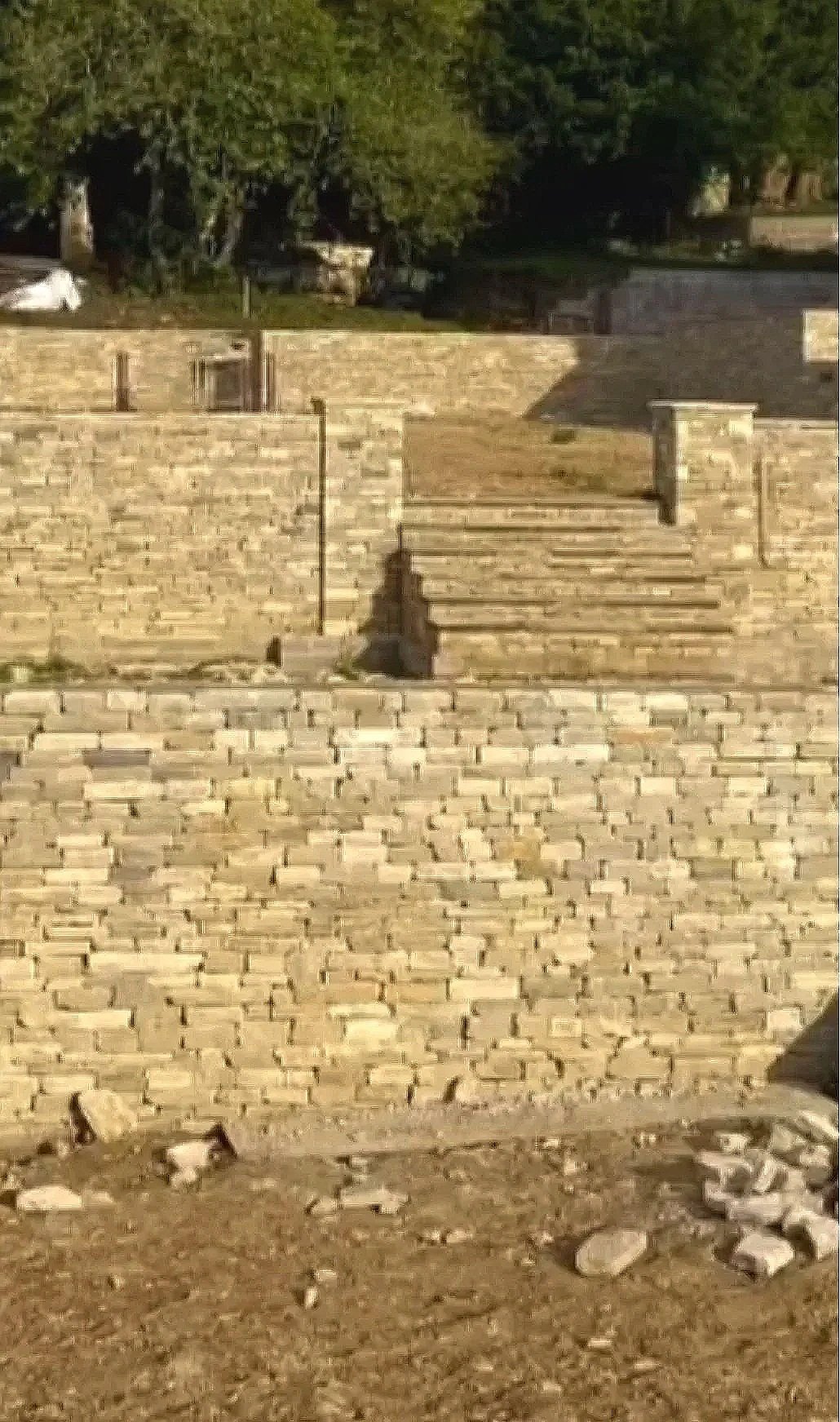 A stone staircase outdoors, leading up to a higher level with a brick retaining wall, surrounded by trees and greenery.