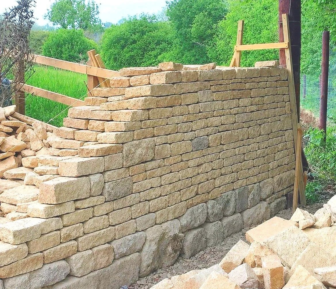 Stacked stone wall under construction with wooden supports in a green outdoor setting.