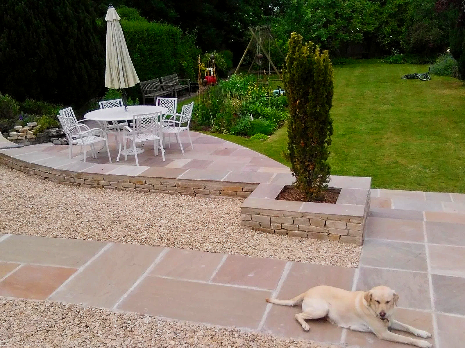 A backyard patio with a white round table and six chairs, a closed outdoor umbrella, a nearby garden with dense green trees, and a grassy lawn. A dog is lying on the patio in the foreground, and a bicycle is on the grass.