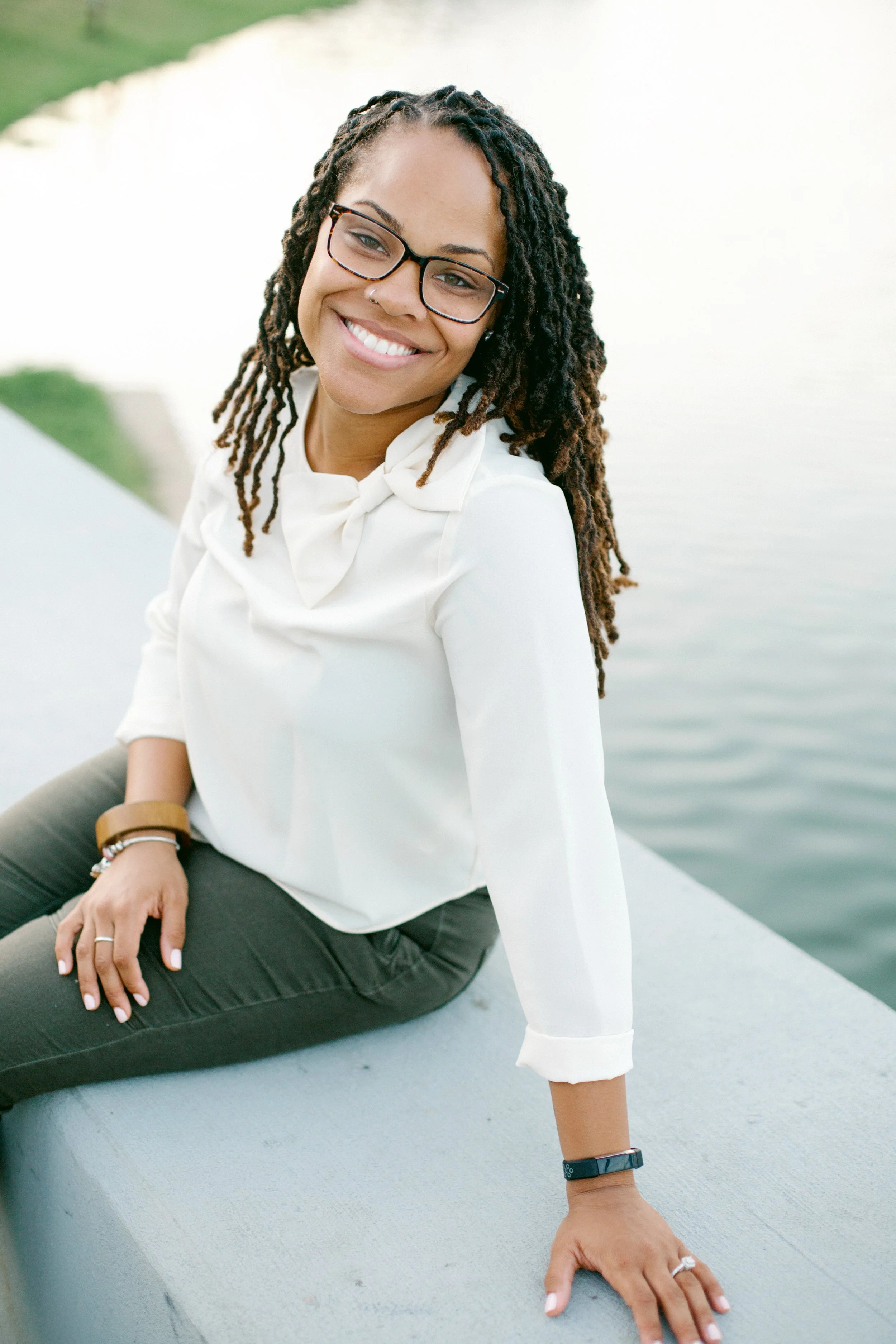 A woman with glasses and dreadlocks smiling outdoors near water, seated on a concrete ledge.