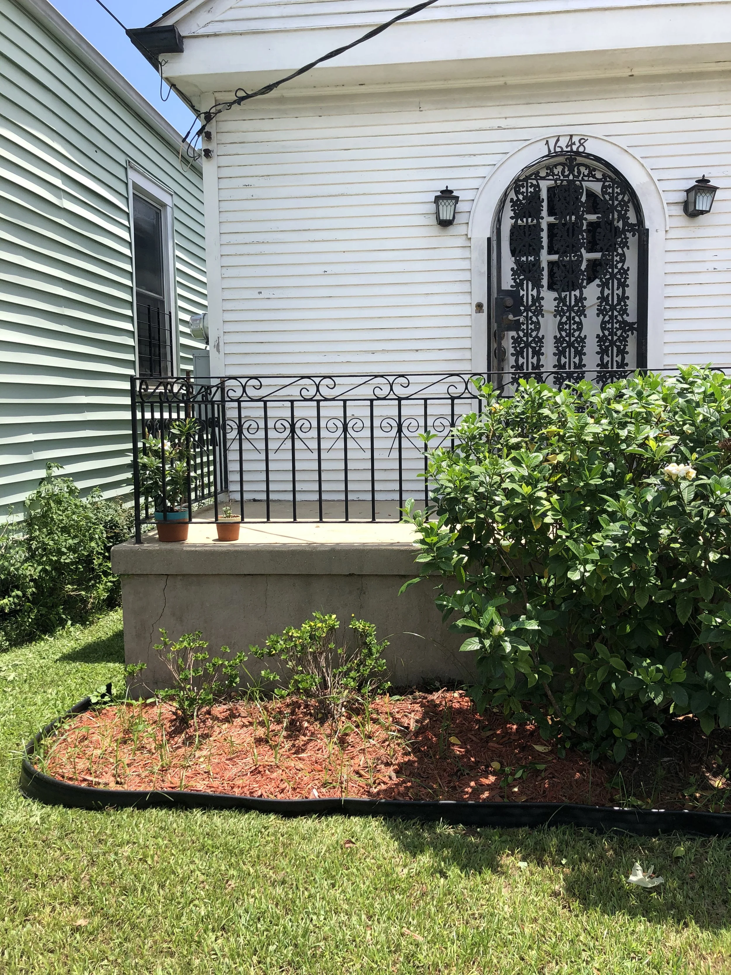 A house entrance featuring a black metal door with intricate patterns, white trim, two outdoor wall lanterns, a small balcony with a black railing, and a yard with a garden bed of plants and bushes.