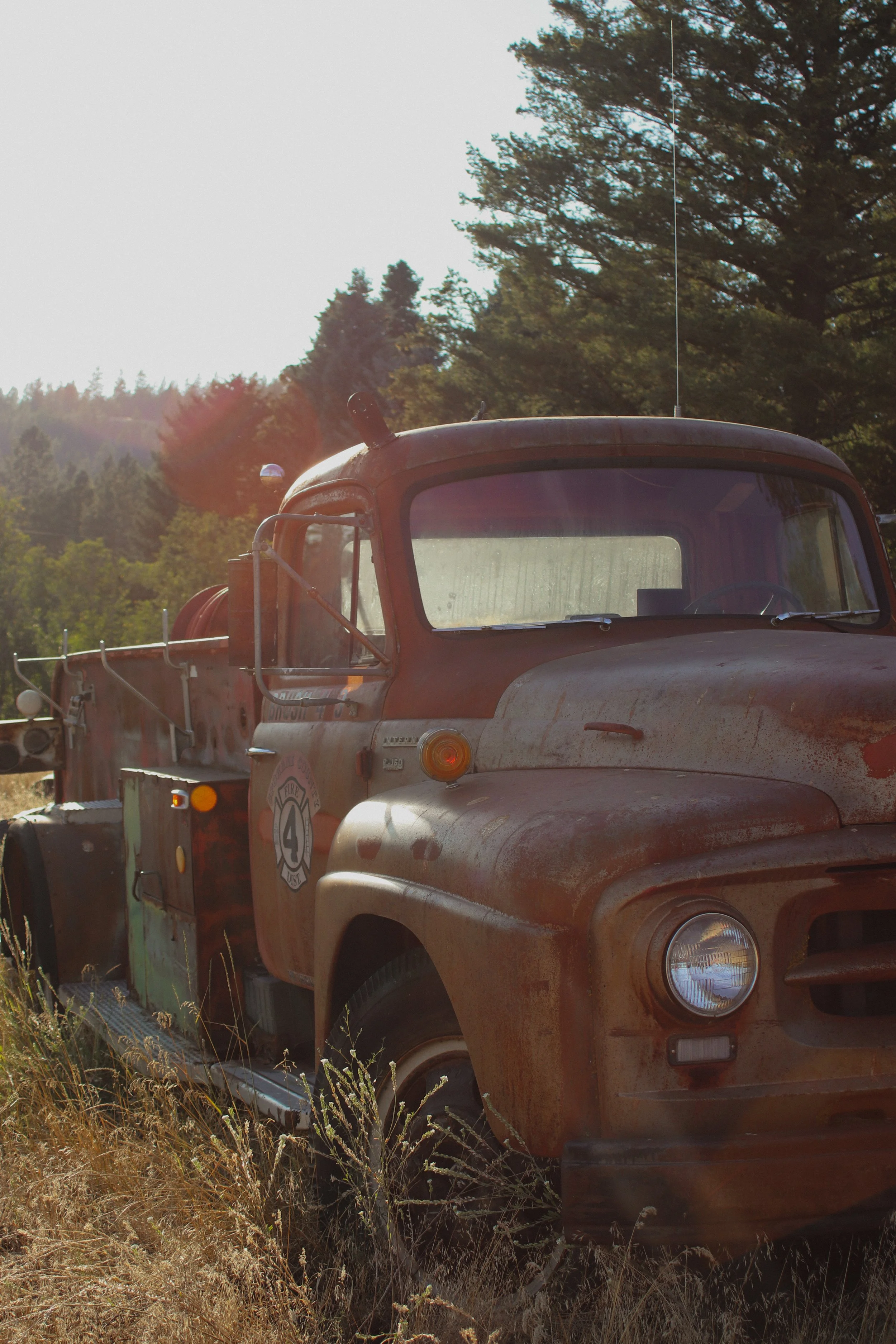 Old rusty fire truck parked in a field with dry grass and trees in the background.