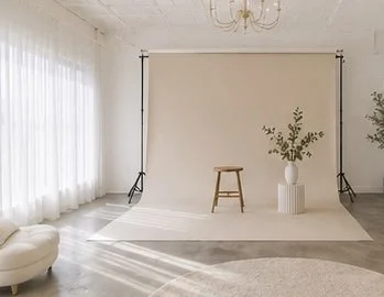 Photo of a photography studio with a beige backdrop, a wooden stool, a white vase with green branches, and a soft rug on the floor.