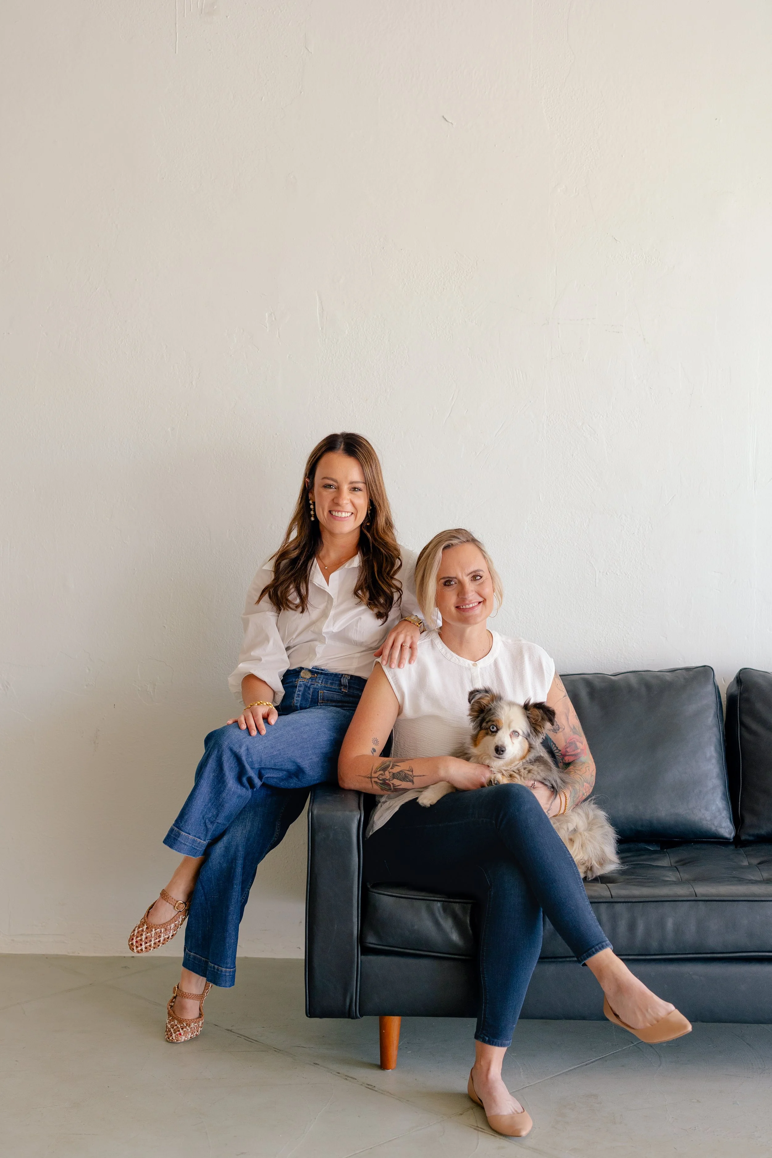 Two women in stylish black outfits pose in front of an exposed brick wall, with one sitting on a gold chair and the other standing beside her.