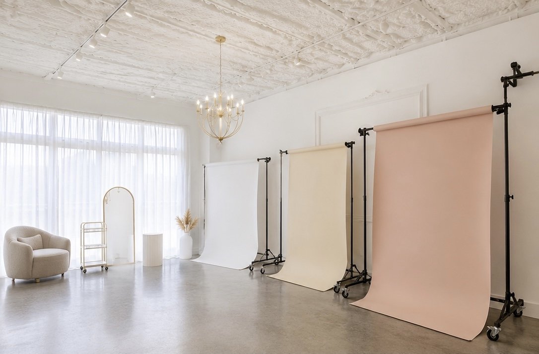 Empty photography studio with three backdrop stands holding white, cream, and blush pink backdrops, a beige armchair, a full-length mirror, a white cylindrical lamp, and a chandelier, with natural light coming through sheer curtains.