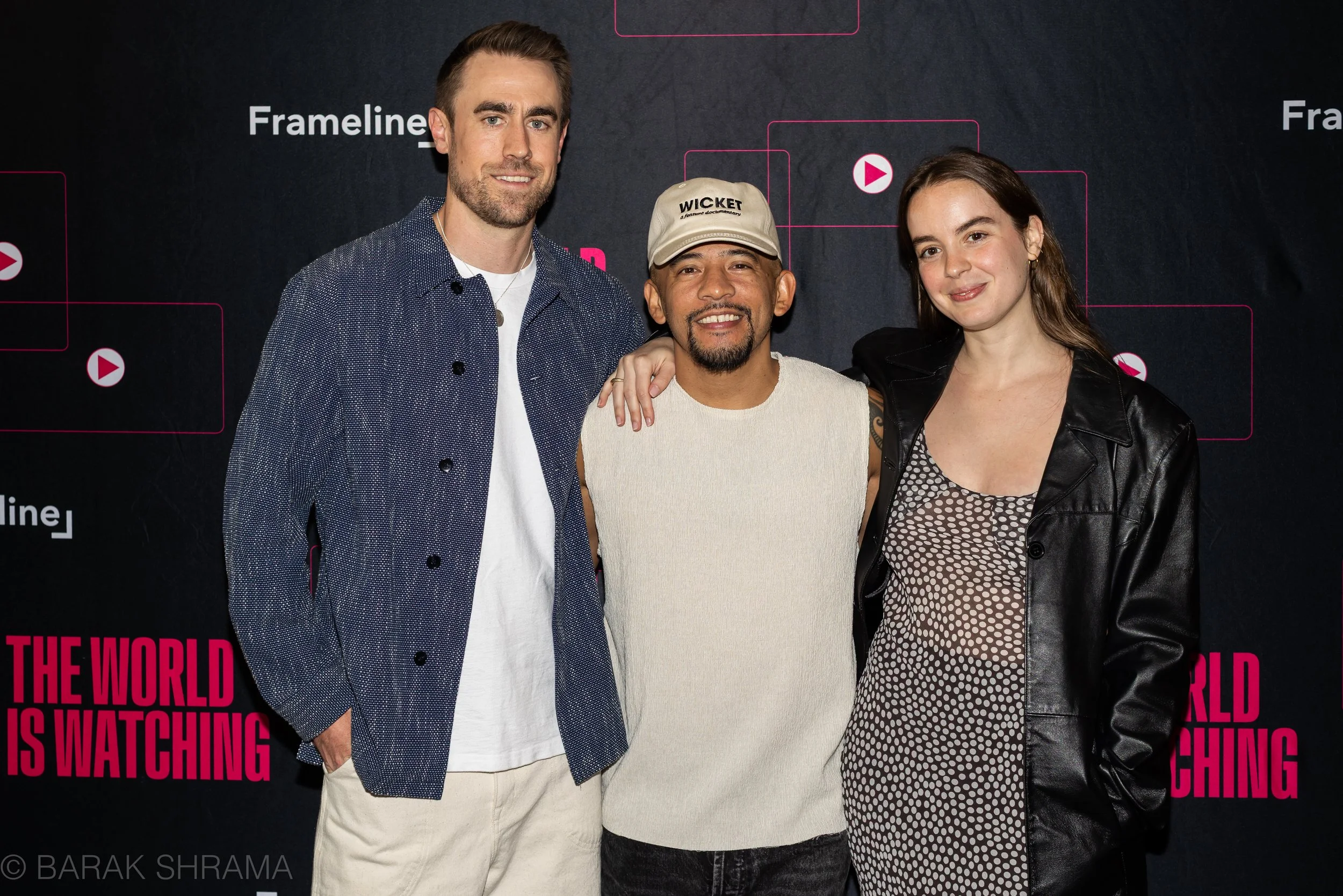 Three people standing together in front of a black background with pink text and graphics. The person on the left is a man with brown hair and a beard, wearing a blue and white patterned shirt. The person in the middle is a man wearing a beige cap with text, a white sleeveless shirt, and has a goatee. The person on the right is a woman with brown hair, wearing a black leather jacket and a polka dot dress. All three are smiling.