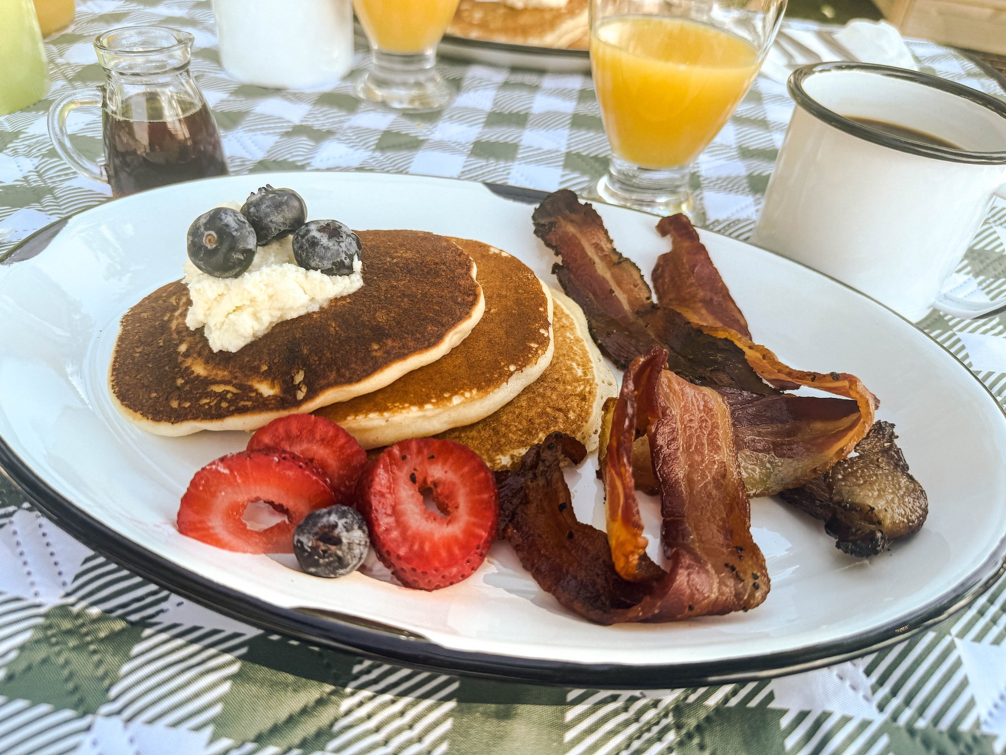 Plate of 3 pancakes topped with whipped cream, three blueberries, next to sliced strawberries and several pieces of bacon