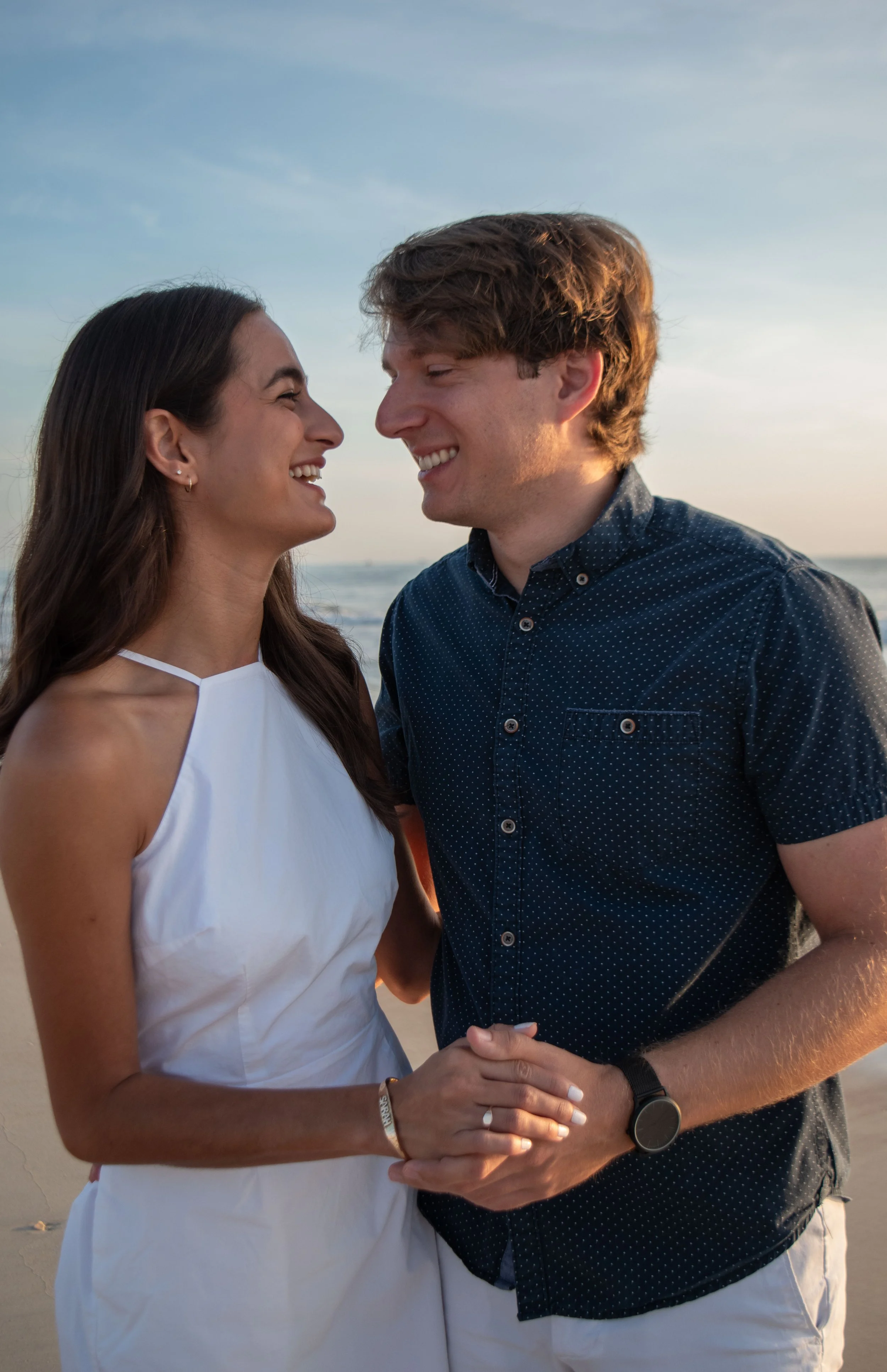 A couple holding hands and smiling at each other on the beach during sunset.
