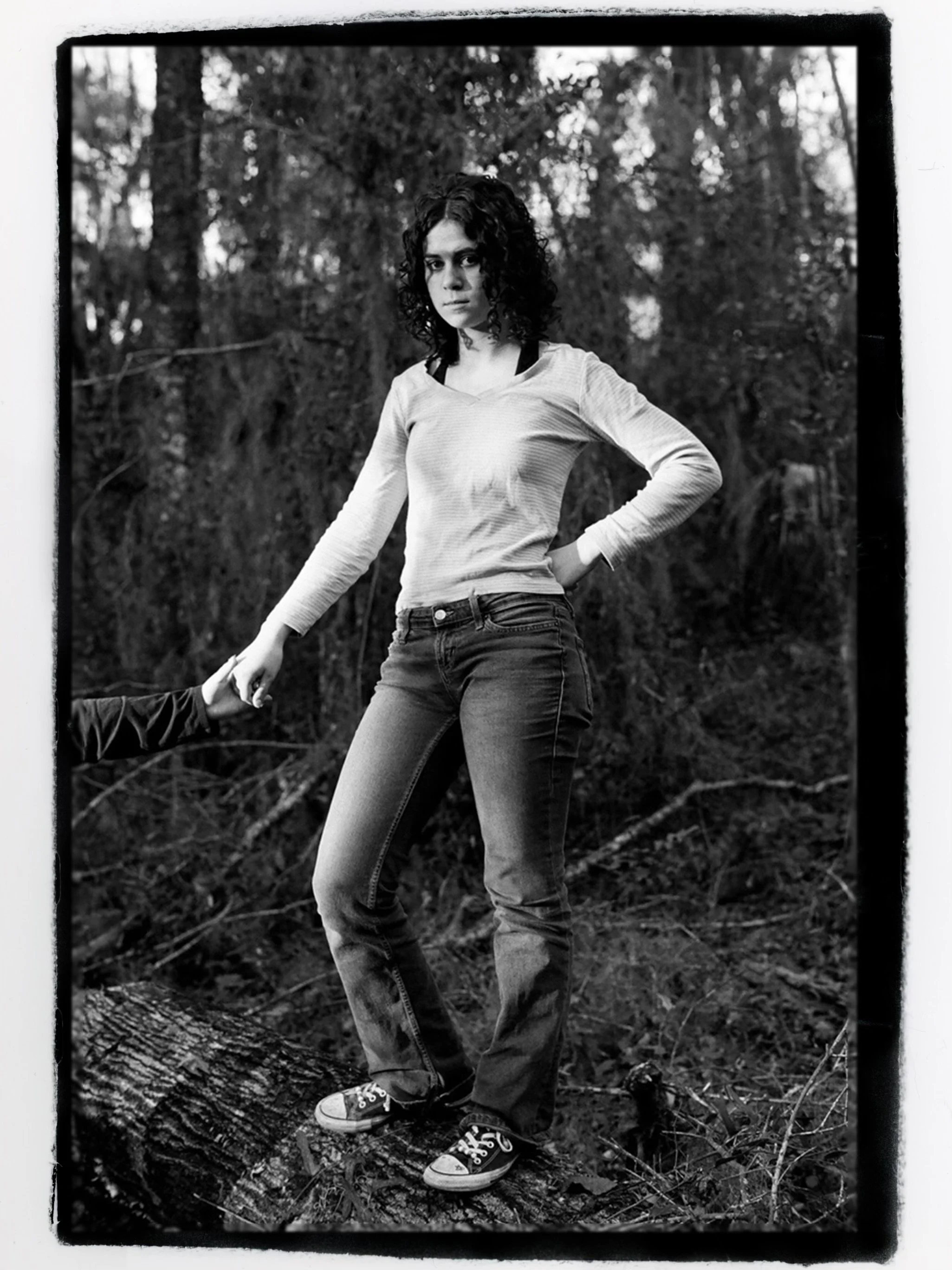 A young woman with curly hair stands outdoors in a wooded area, holding someone's hand, wearing a long sleeve shirt, jeans, and sneakers, with a serious expression.