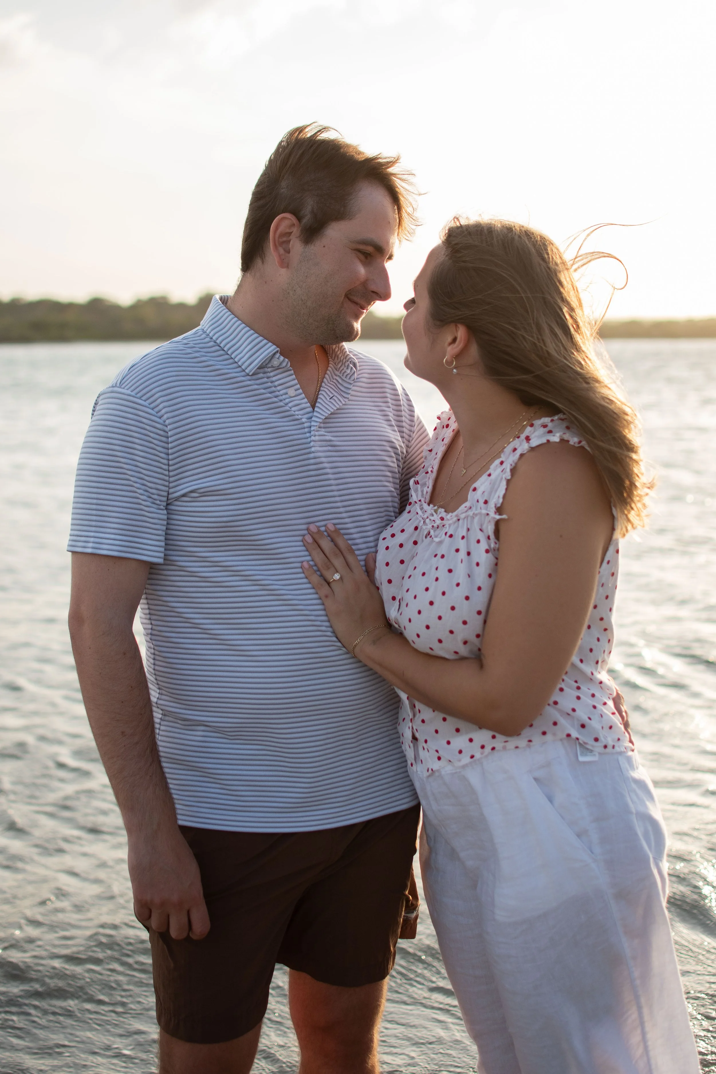 A man and woman standing close together by the water at sunset, smiling at each other, with the woman touching the man's chest.