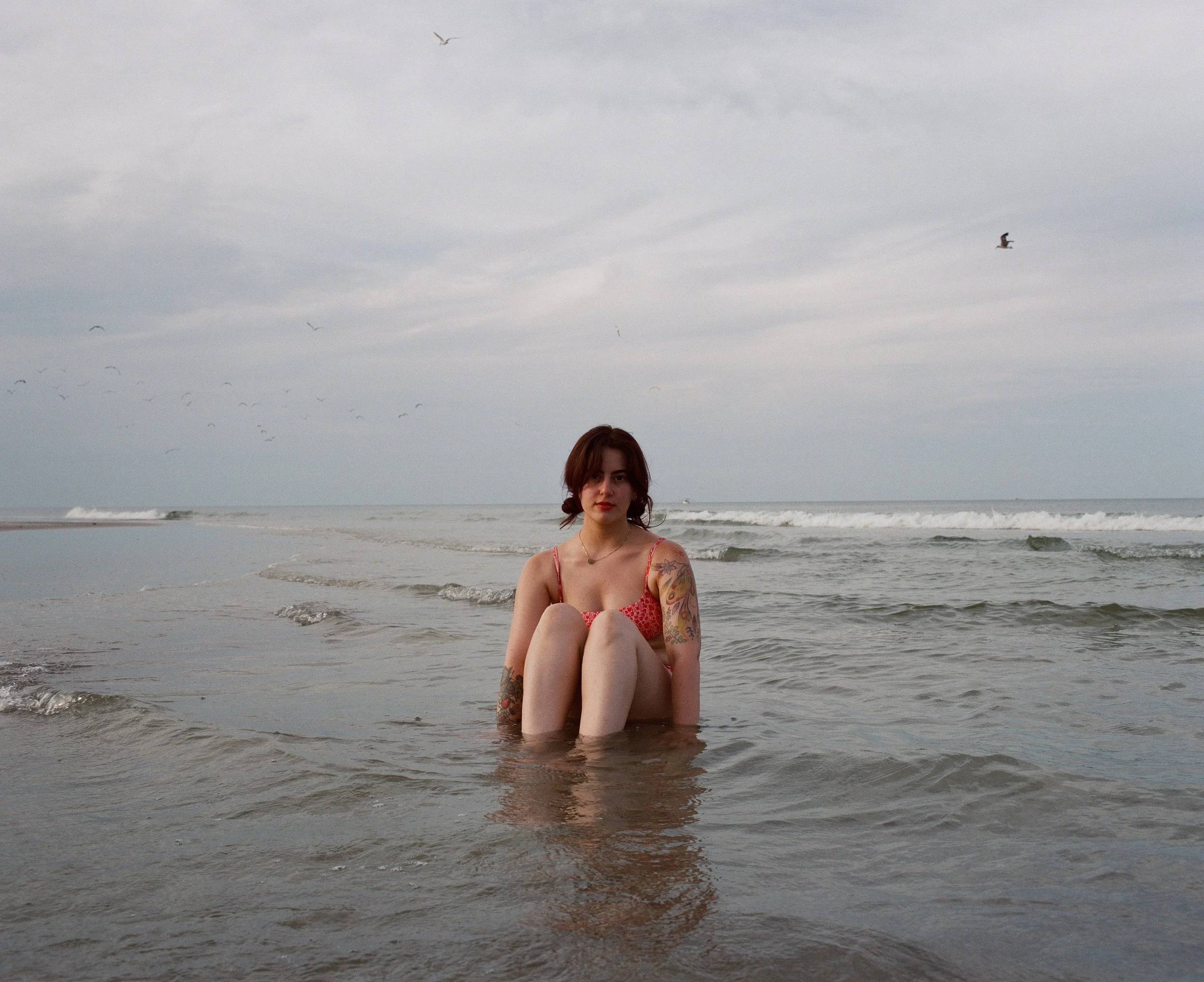 A woman with dark red hair and tattoos on her arm and shoulder, sitting in shallow water at the beach, wearing a red patterned swimsuit, with an overcast sky and seagulls flying overhead.
